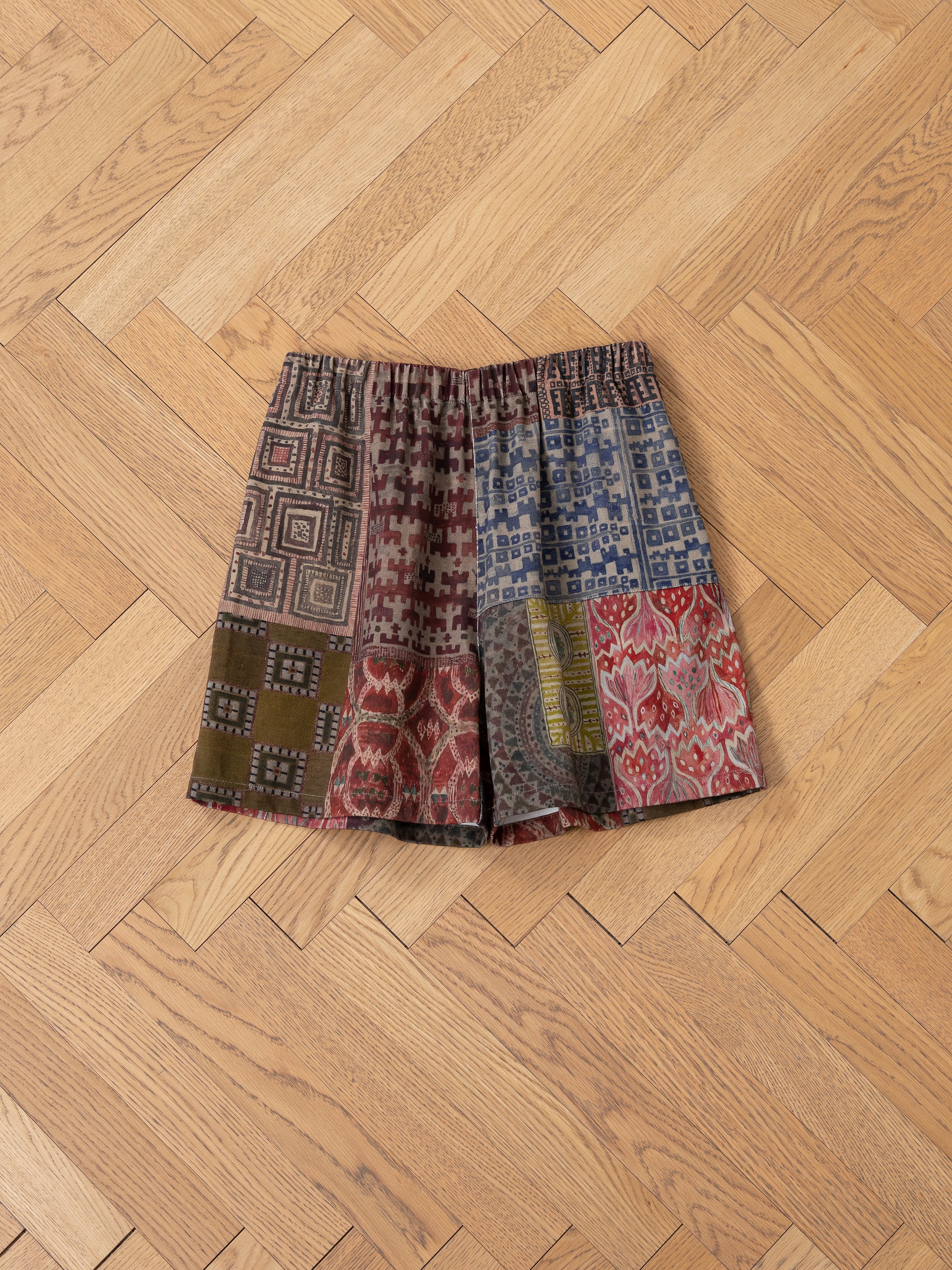 A pair of FOUND's Watercolor Mosaic Work Shorts, unisex with a relaxed fit and featuring vibrant geometric and abstract patterns in a patchwork design, laid flat on a wooden parquet floor.