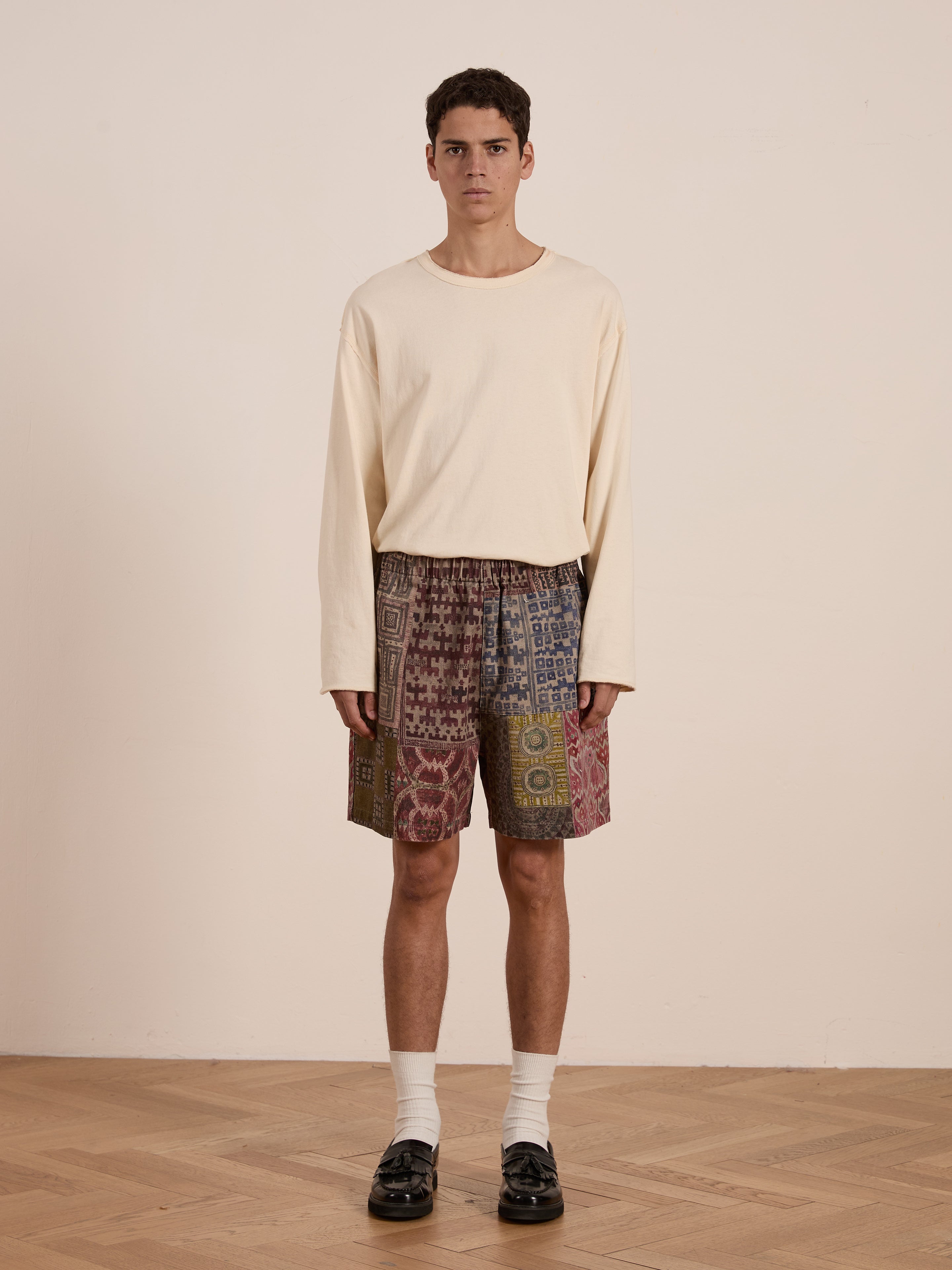 A person in a long-sleeve beige shirt and FOUND's Watercolor Mosaic Work Shorts with vintage-inspired patterns stands on a wooden floor, complemented by white socks and black shoes, creating a unisex relaxed fit against the plain light background.