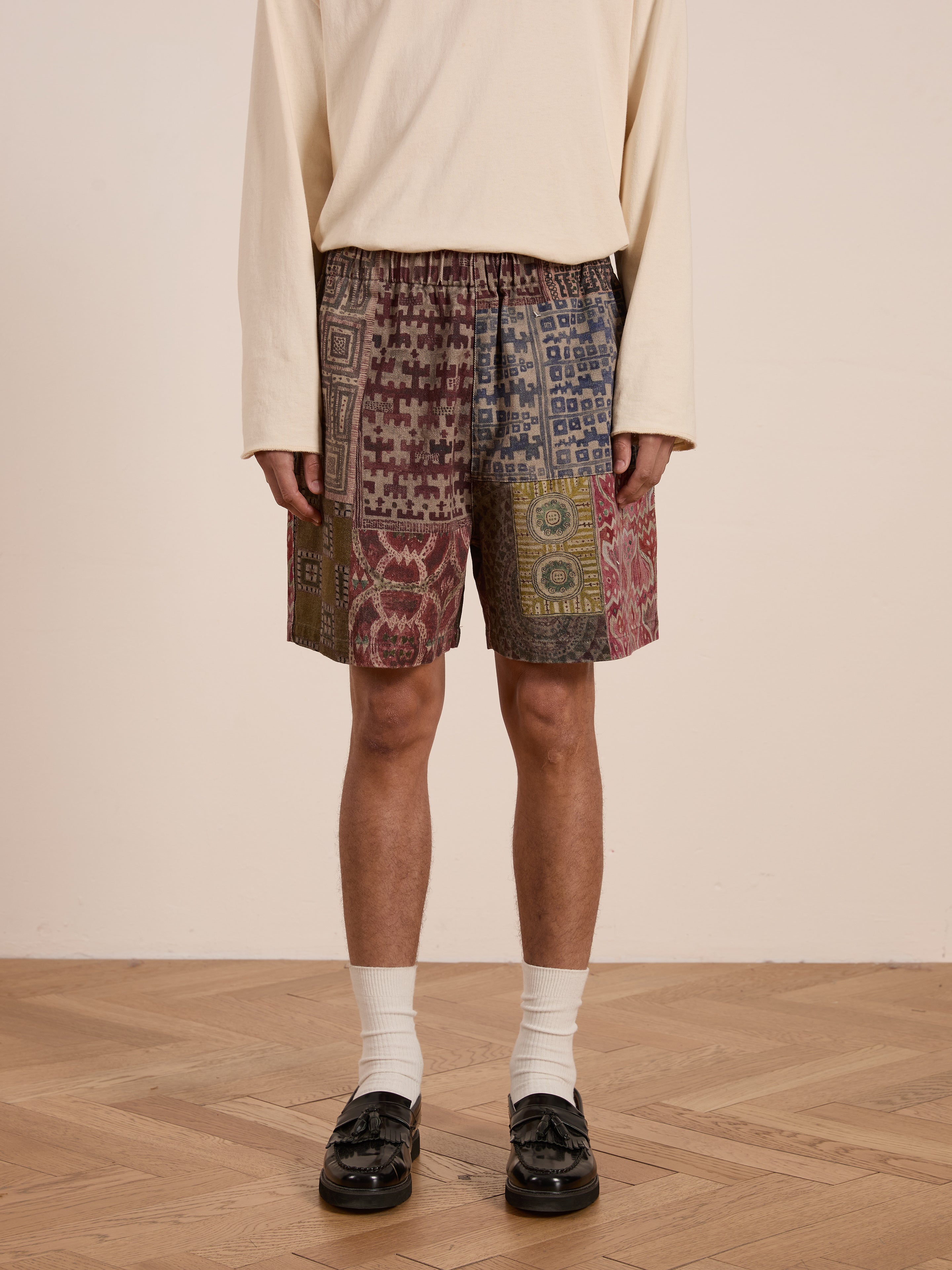 Dressed in a cream long-sleeve shirt and FOUND's Watercolor Mosaic Work Shorts with vintage-inspired patterns, the person completes their unisex relaxed fit with white socks and black loafers, standing on a wooden floor.