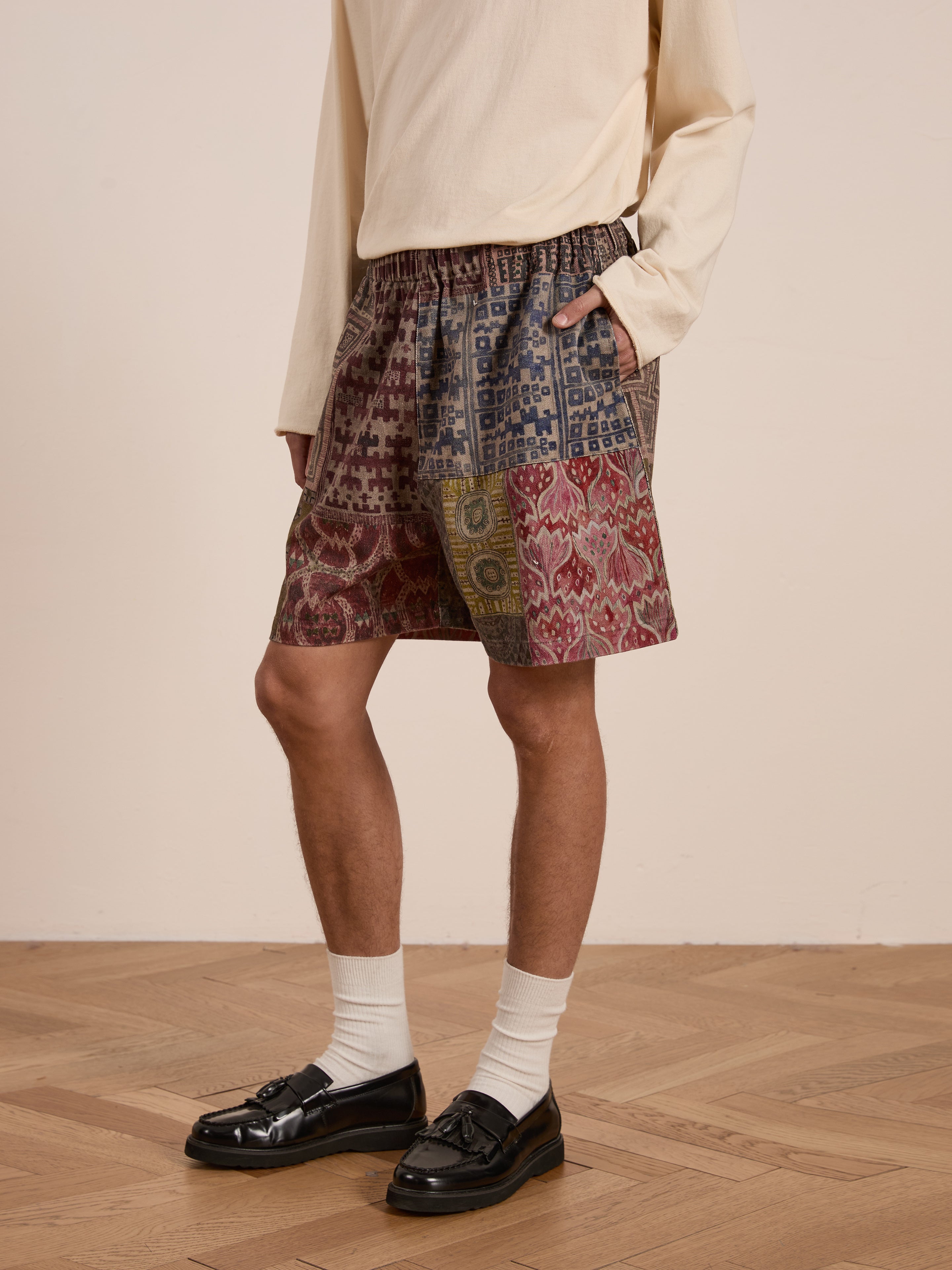 A person in a long-sleeve beige shirt and FOUND's Watercolor Mosaic Work Shorts, featuring vintage-inspired patterns, stands on a wooden floor. The outfit is completed with white socks and black loafers for a unisex relaxed fit look.