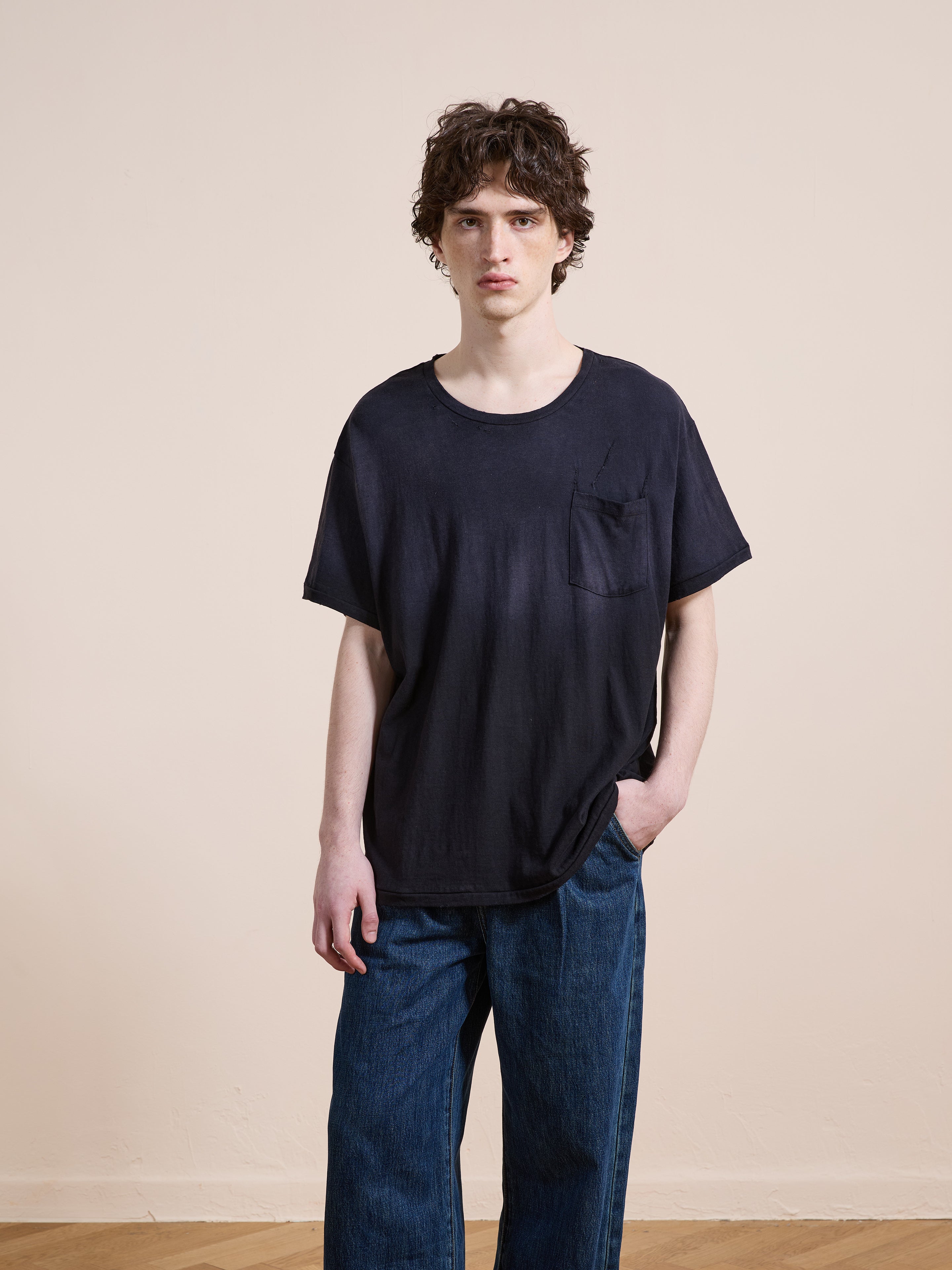 A person with wavy brown hair wears the FOUND Weathered Pocket Tee—a relaxed fit black t-shirt with a chest pocket—paired with blue jeans, standing indoors against a plain light-colored wall.