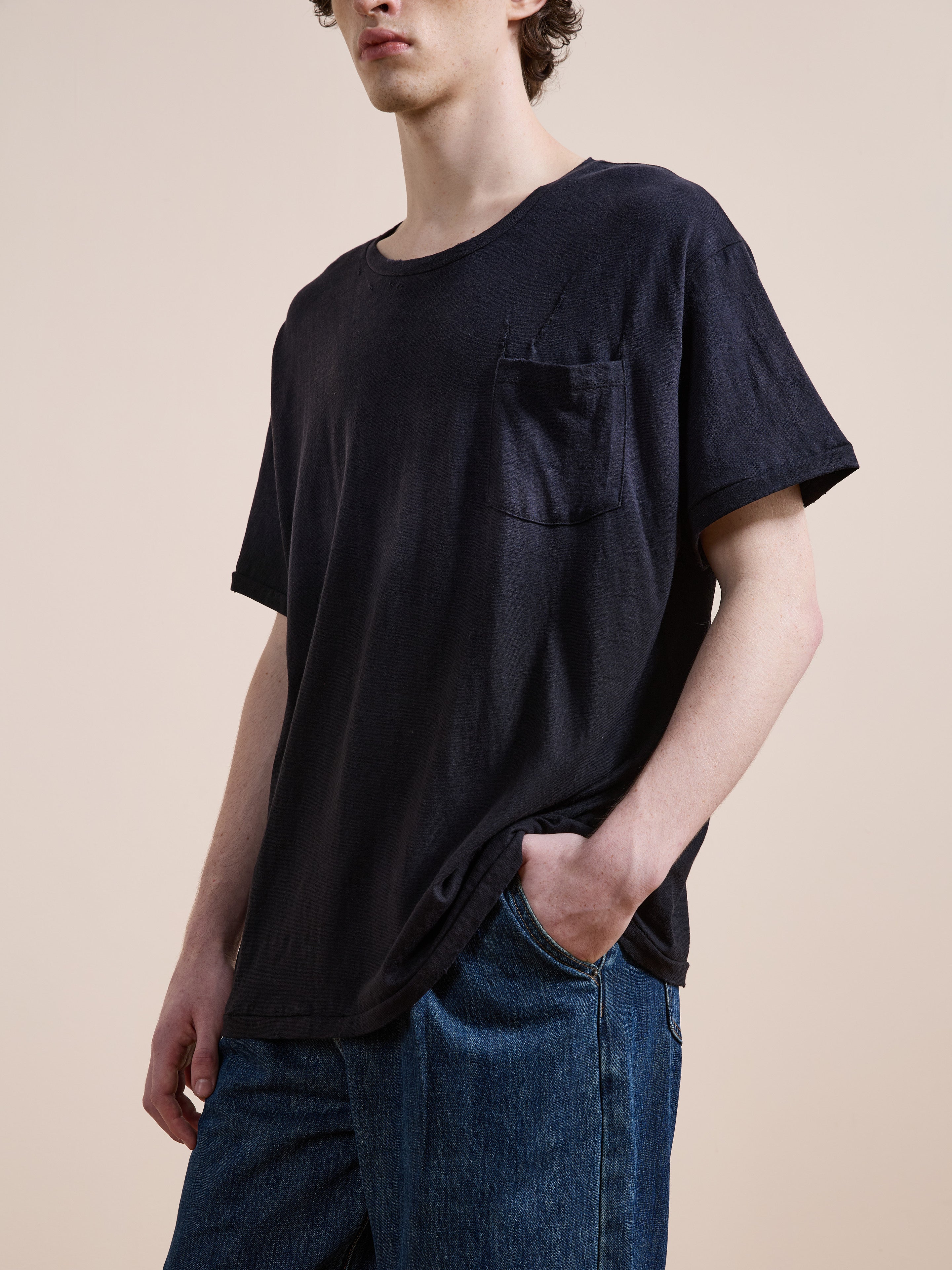 Someone stands against a plain background wearing the FOUND Weathered Pocket Tee—a unisex, relaxed fit black T-shirt with a chest pocket—paired with blue jeans and one hand in their pocket.