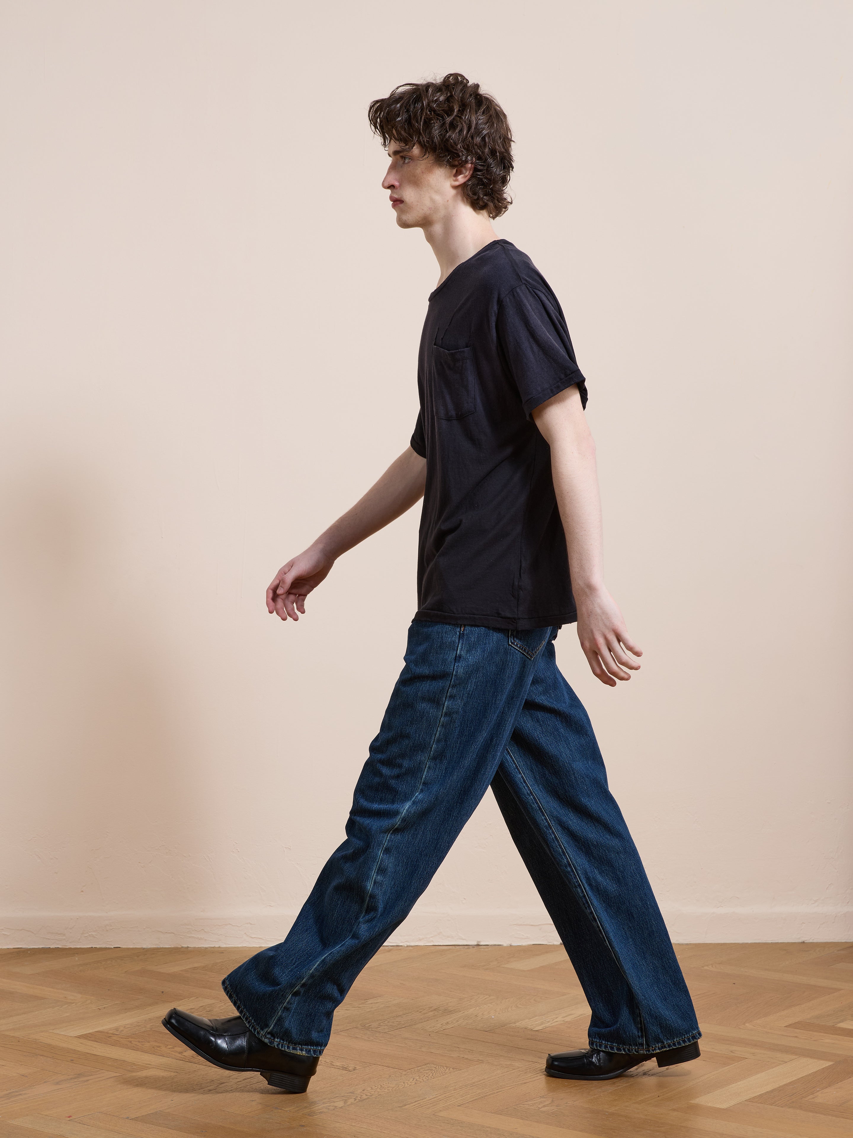 A person with curly hair walks indoors on a wooden floor, wearing the FOUND Weathered Pocket Tee in unisex relaxed fit, paired with loose blue jeans and black shoes against a plain beige wall.