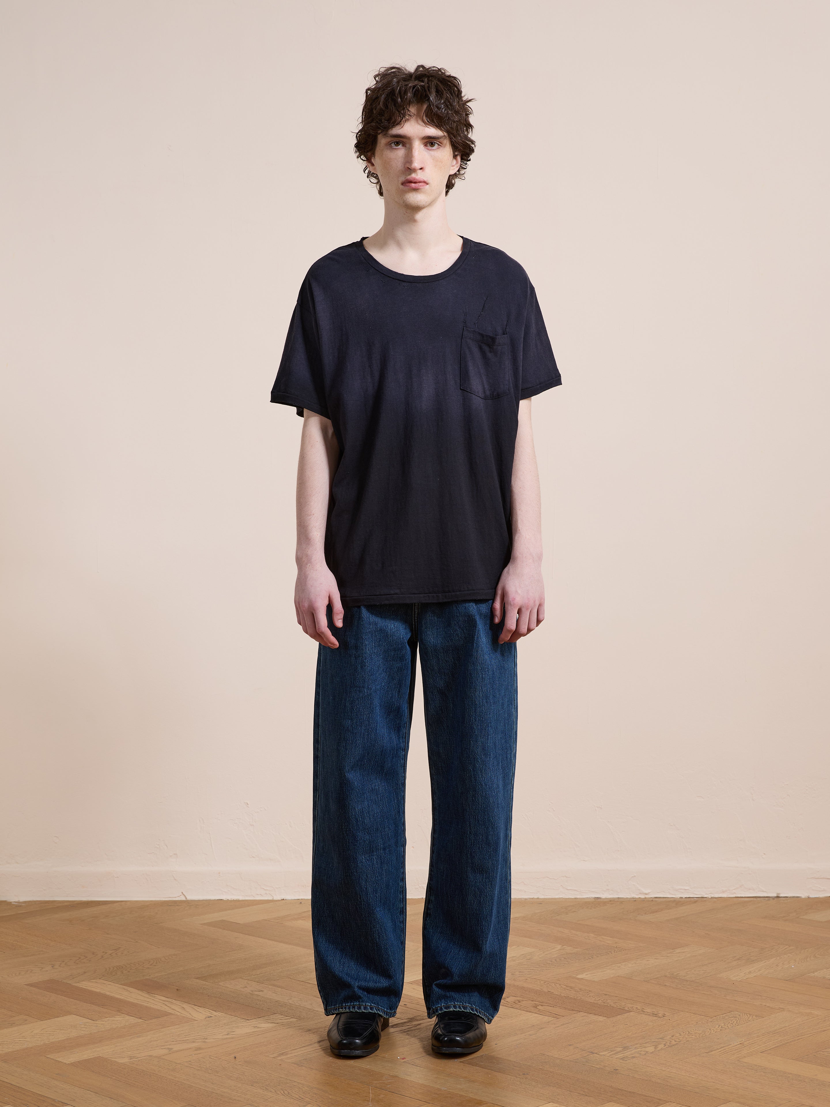 A person stands indoors on a wooden floor wearing FOUND’s Weathered Pocket Tee in black with a unisex relaxed fit, wide blue jeans, and black shoes, set against a plain beige wall.