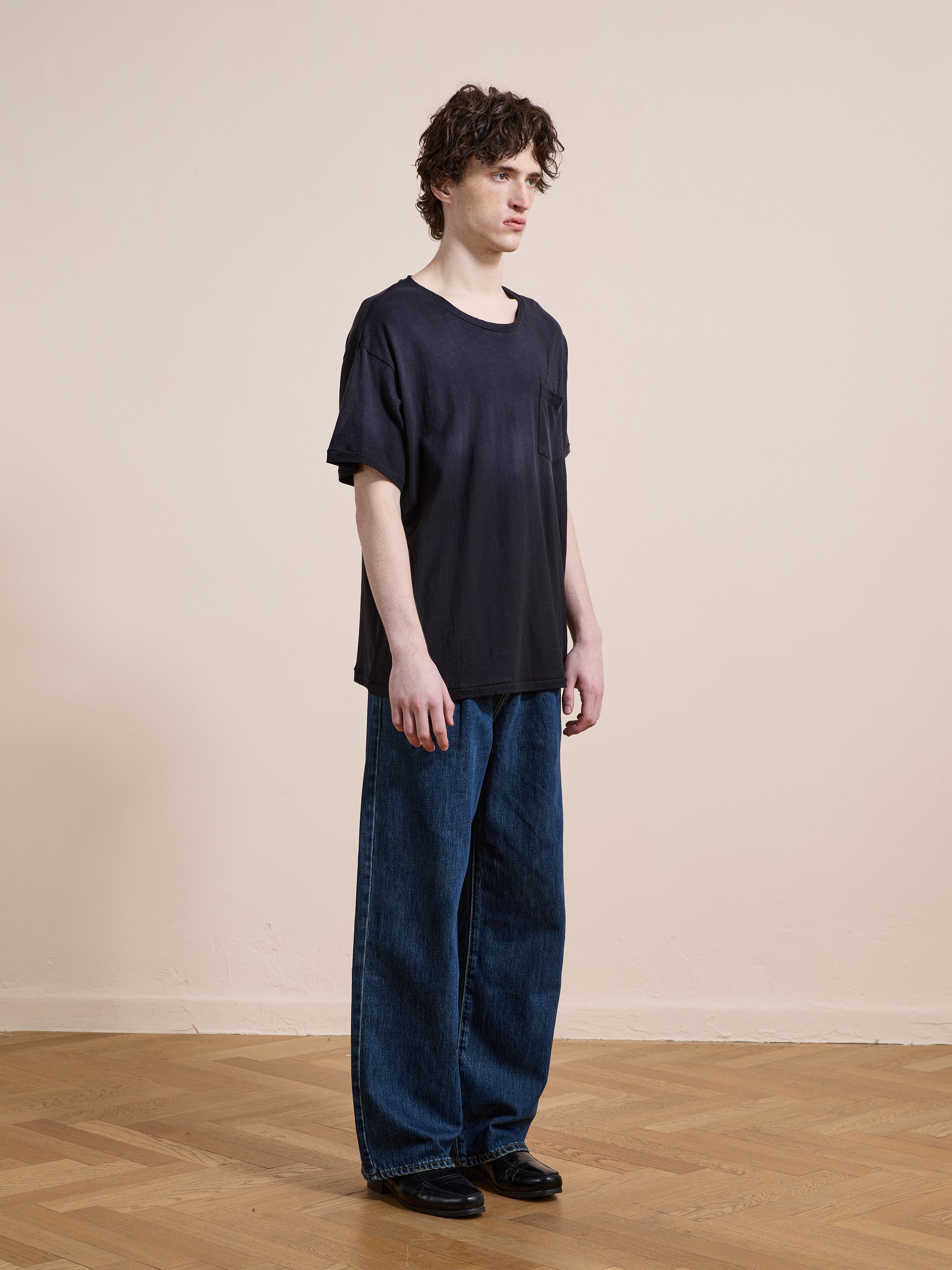 A person stands indoors on a wooden floor, facing slightly to the side against a plain beige wall, wearing FOUND's Weathered Pocket Tee in black, paired with wide-leg blue jeans and black shoes.