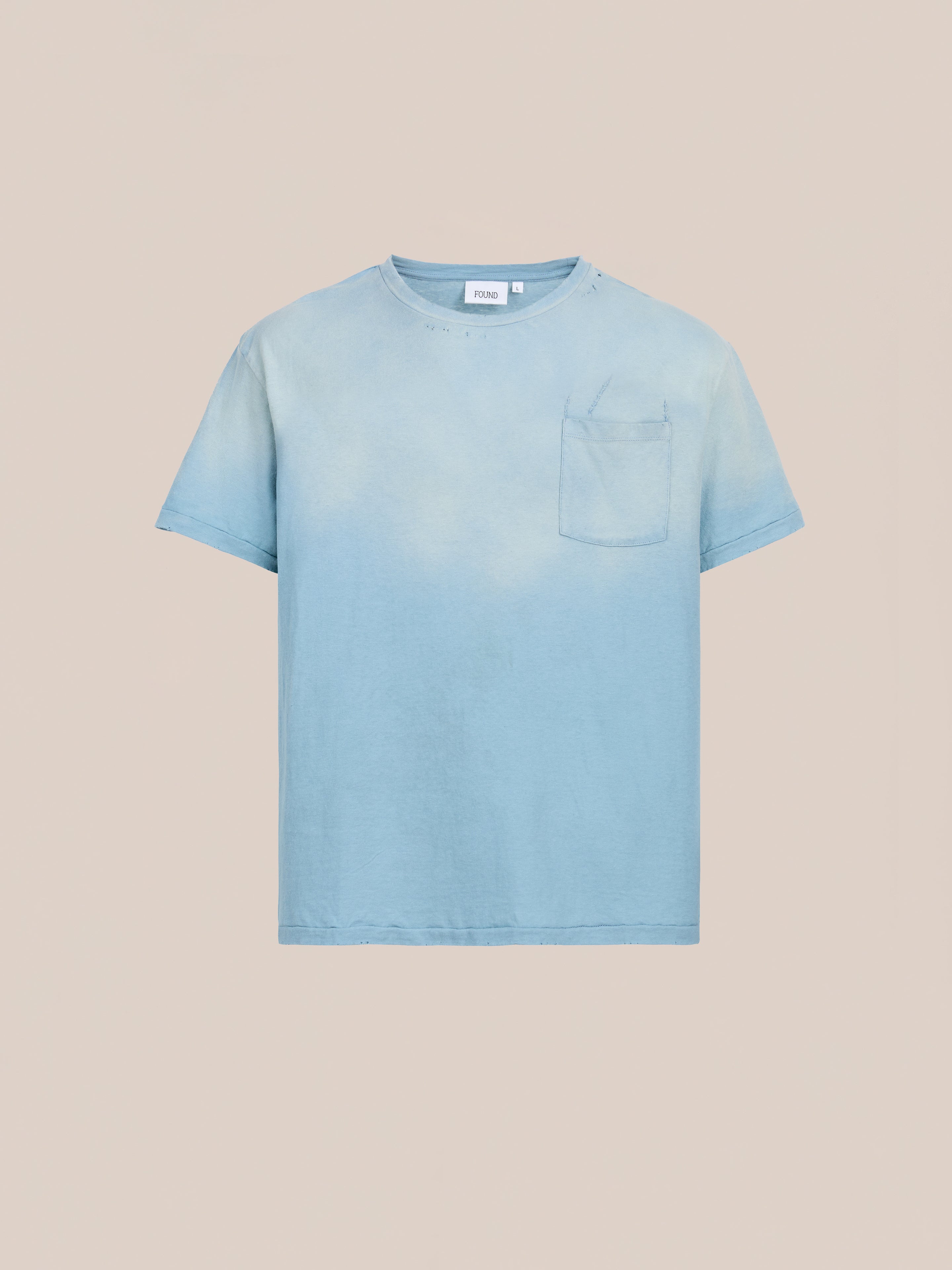 The FOUND Weathered Pocket Tee is a light blue unisex crew neck with short sleeves, a chest pocket, and a relaxed fit, shown against a plain beige background.