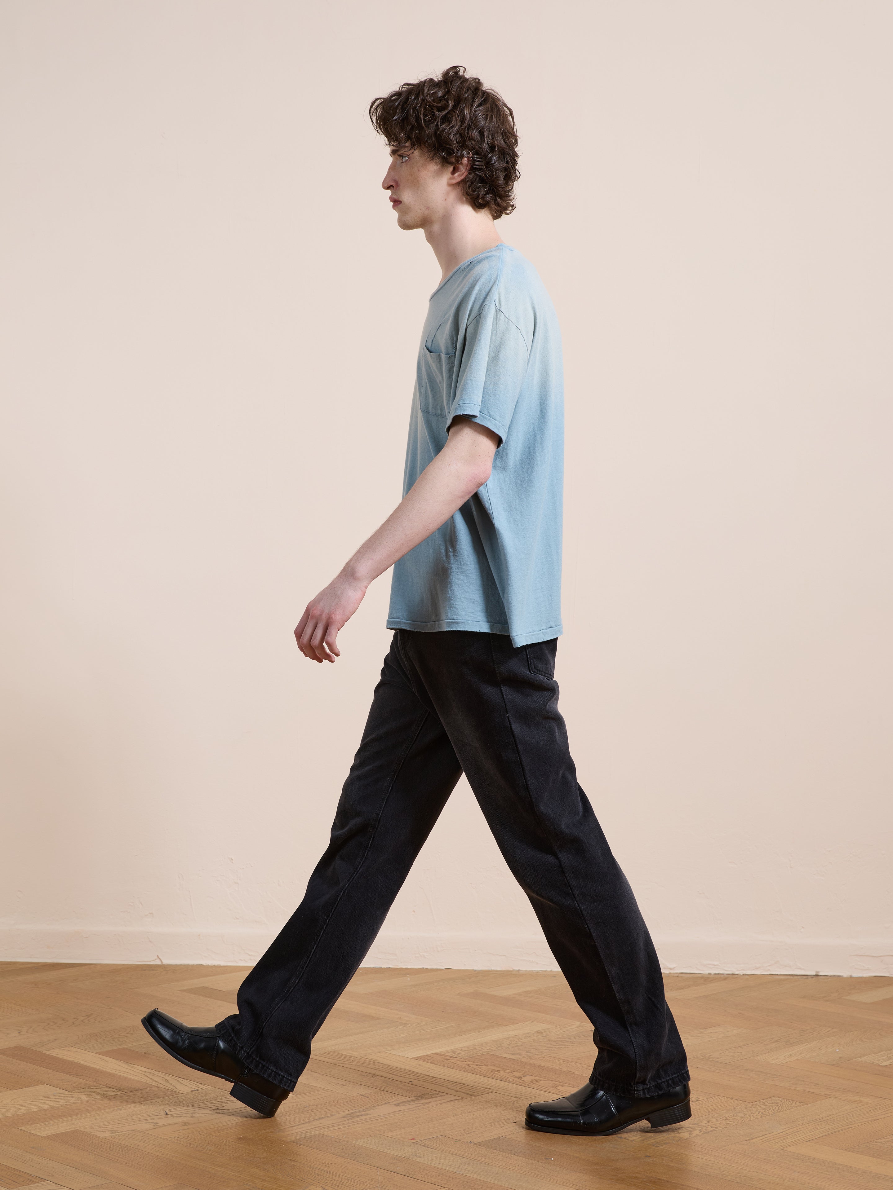 A person with curly hair walks indoors, wearing the FOUND Weathered Pocket Tee in light blue with a unisex relaxed fit, paired with black pants and black shoes on a wooden floor against a plain beige wall.