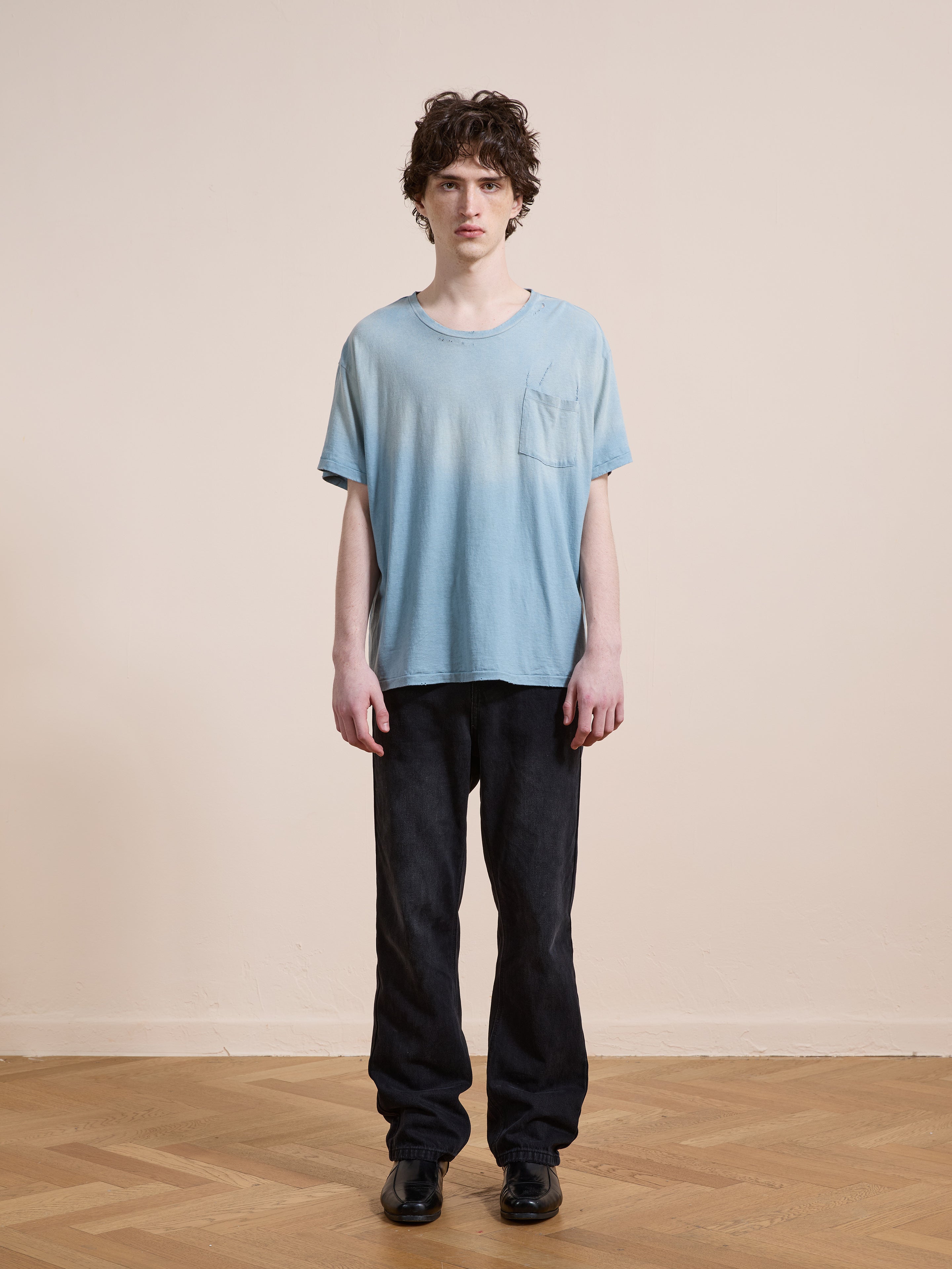 A person stands indoors on a wooden floor, wearing the FOUND Weathered Pocket Tee in light blue, paired with black pants and black shoes, against a plain beige wall.