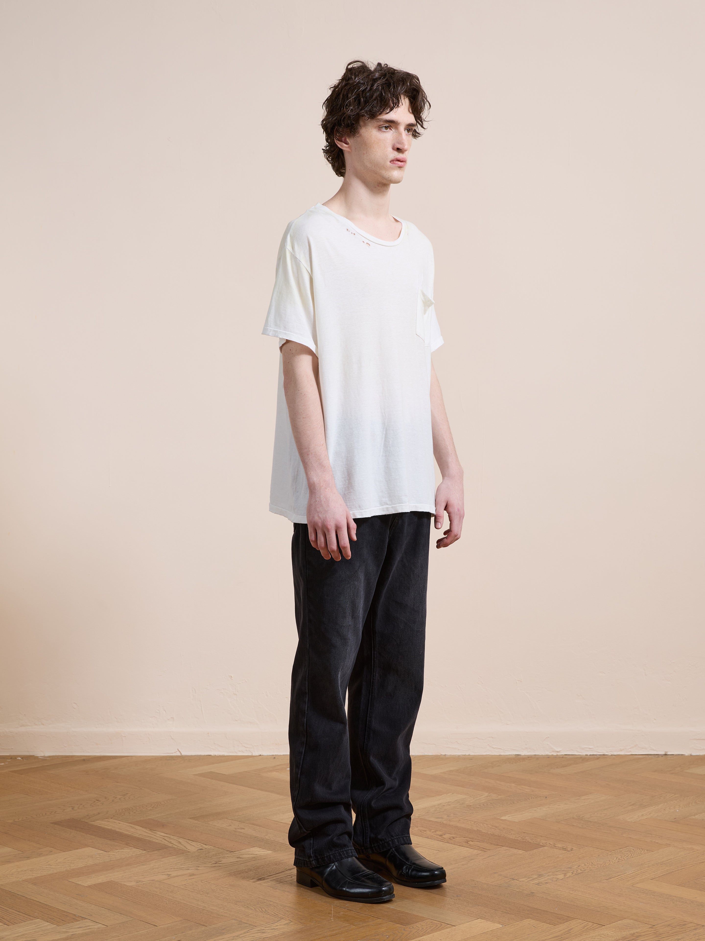 A man stands indoors on a wooden floor against a beige wall, wearing FOUND's Weathered Pocket Tee in white, paired with black pants and black shoes.