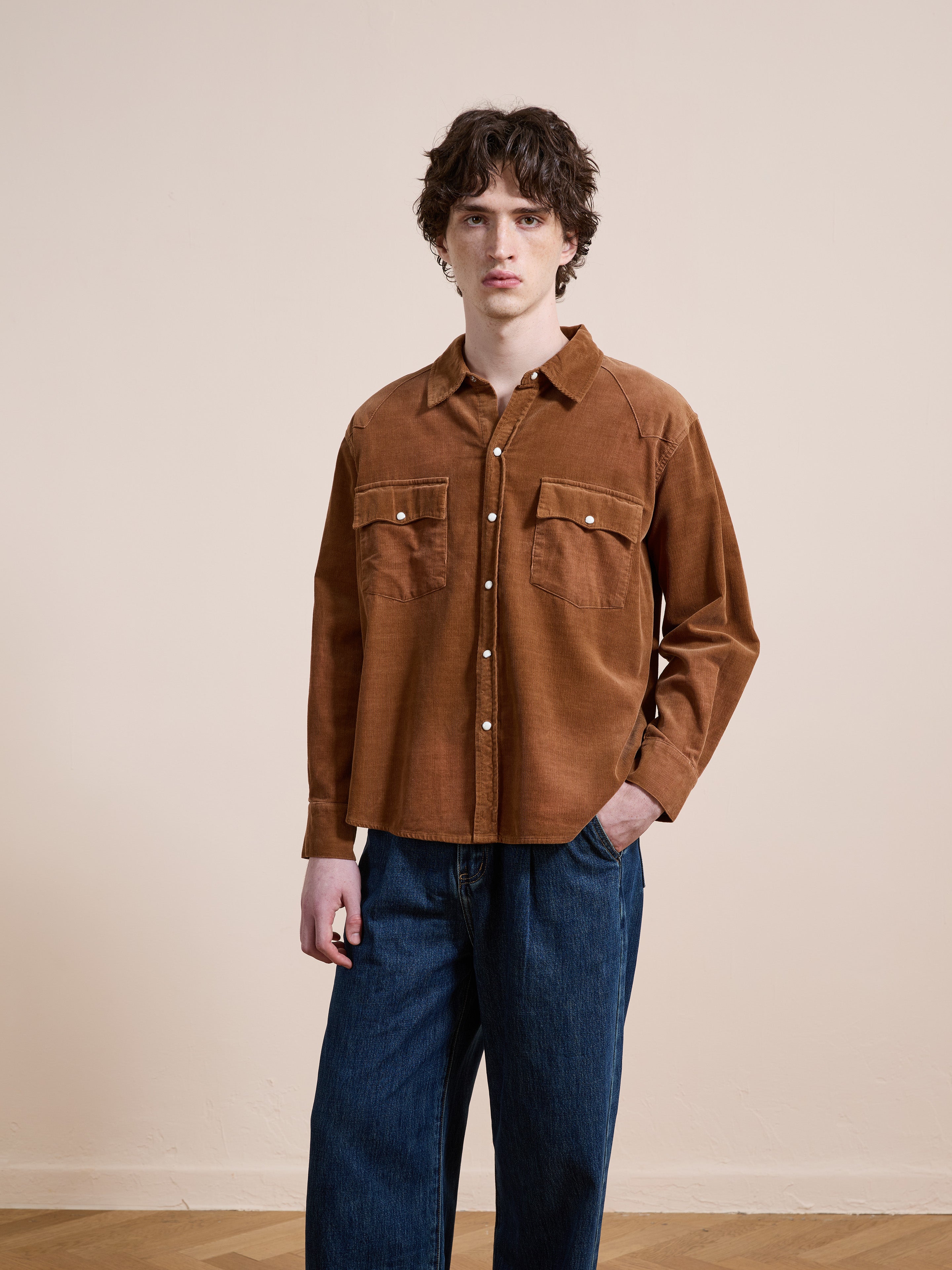 Indoors, a person wears the FOUND Western Corduroy Shirt—a brown unisex relaxed fit button-up with two chest pockets—paired with blue jeans, one hand in their pocket.