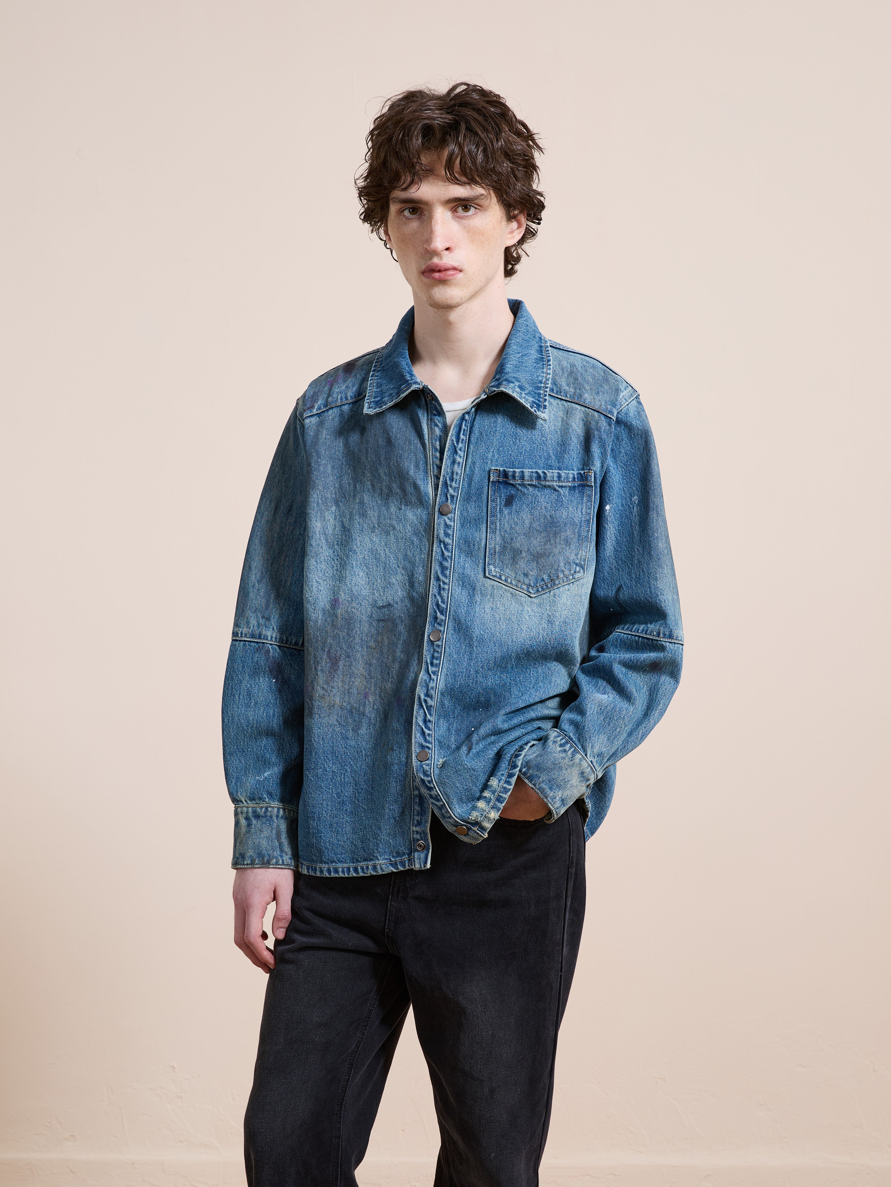 A young person with curly hair wears the FOUND Western Denim Painter Shirt styled as a jacket with black pants, standing against a plain beige background and looking at the camera.