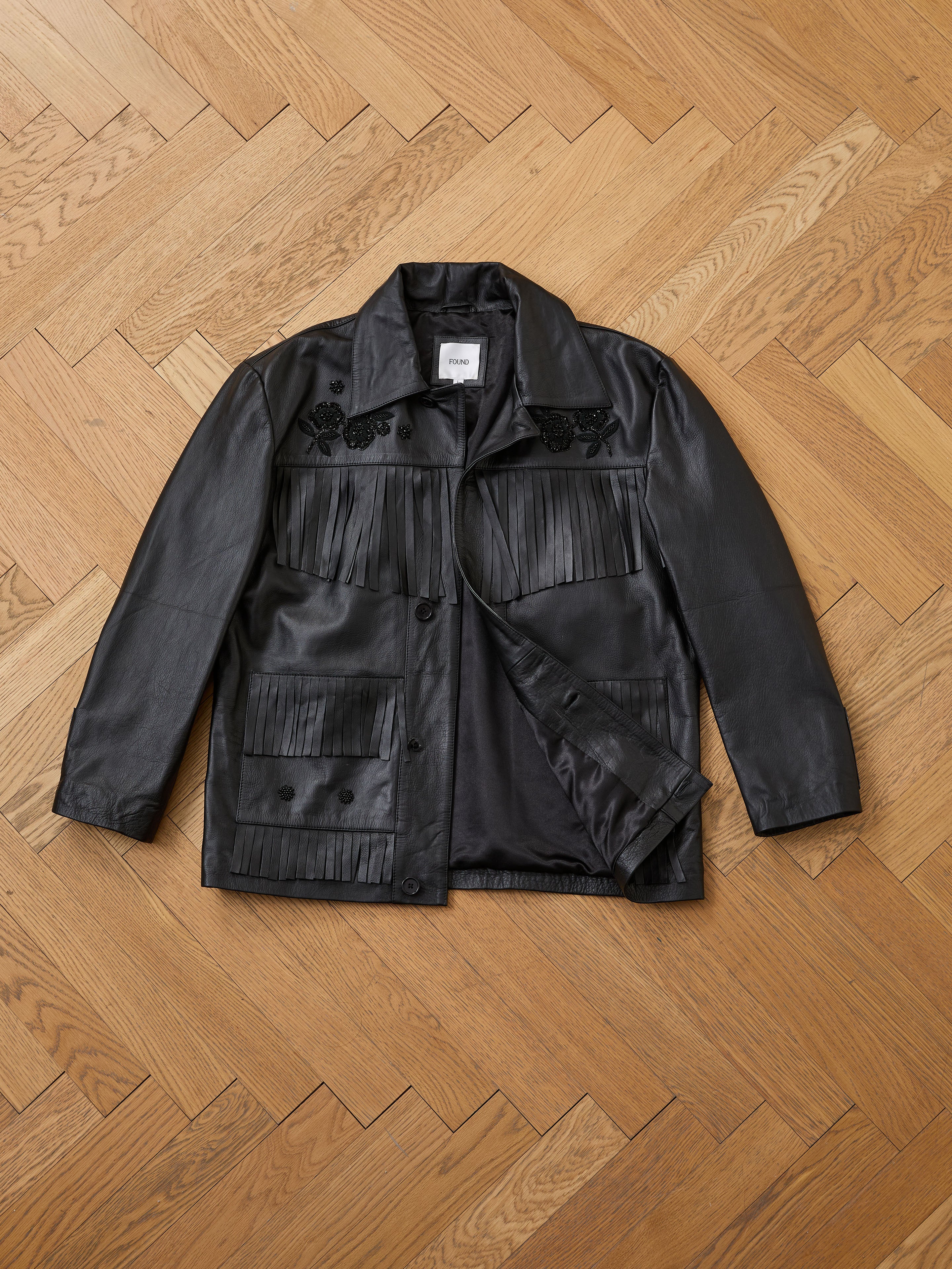 The FOUND Hand Embroidered Leather Fringe Jacket, made from genuine cowhide with fringe accents and intricate embroidery, is shown laid flat on a light herringbone wood floor.