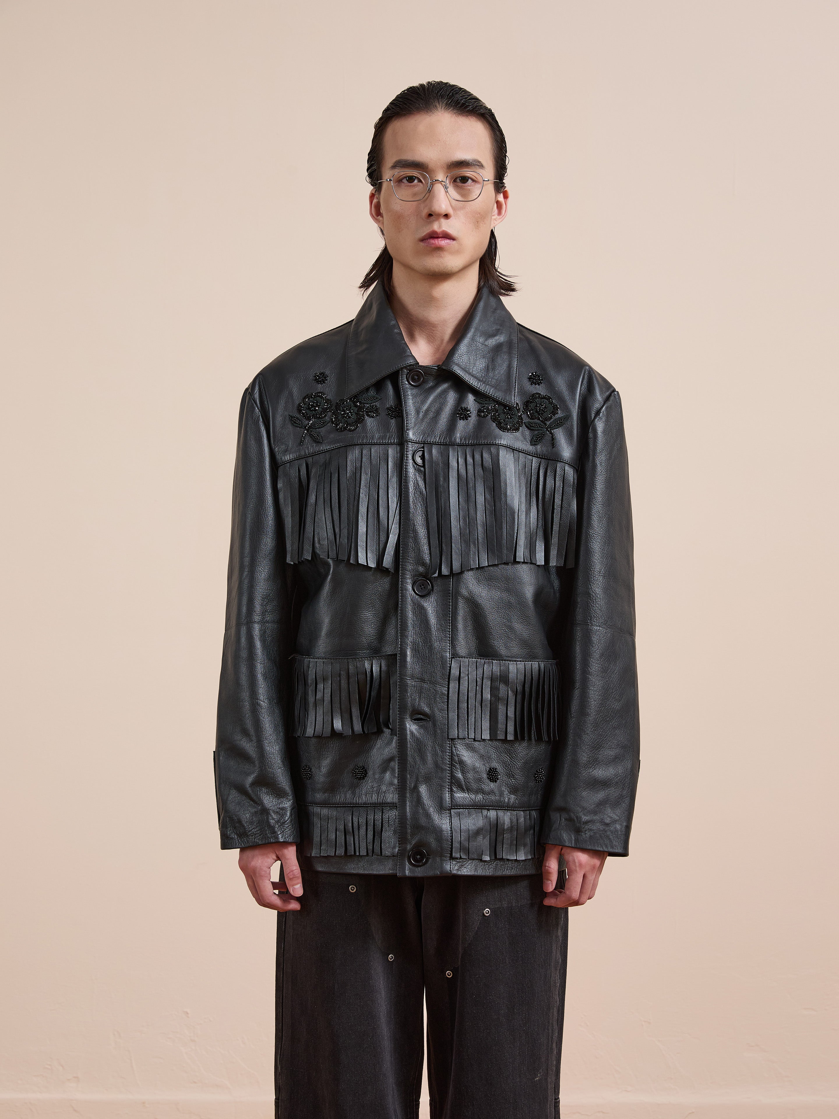 A person stands against a plain background wearing the FOUND Hand Embroidered Leather Fringe Jacket, styled with dark pants and glasses.