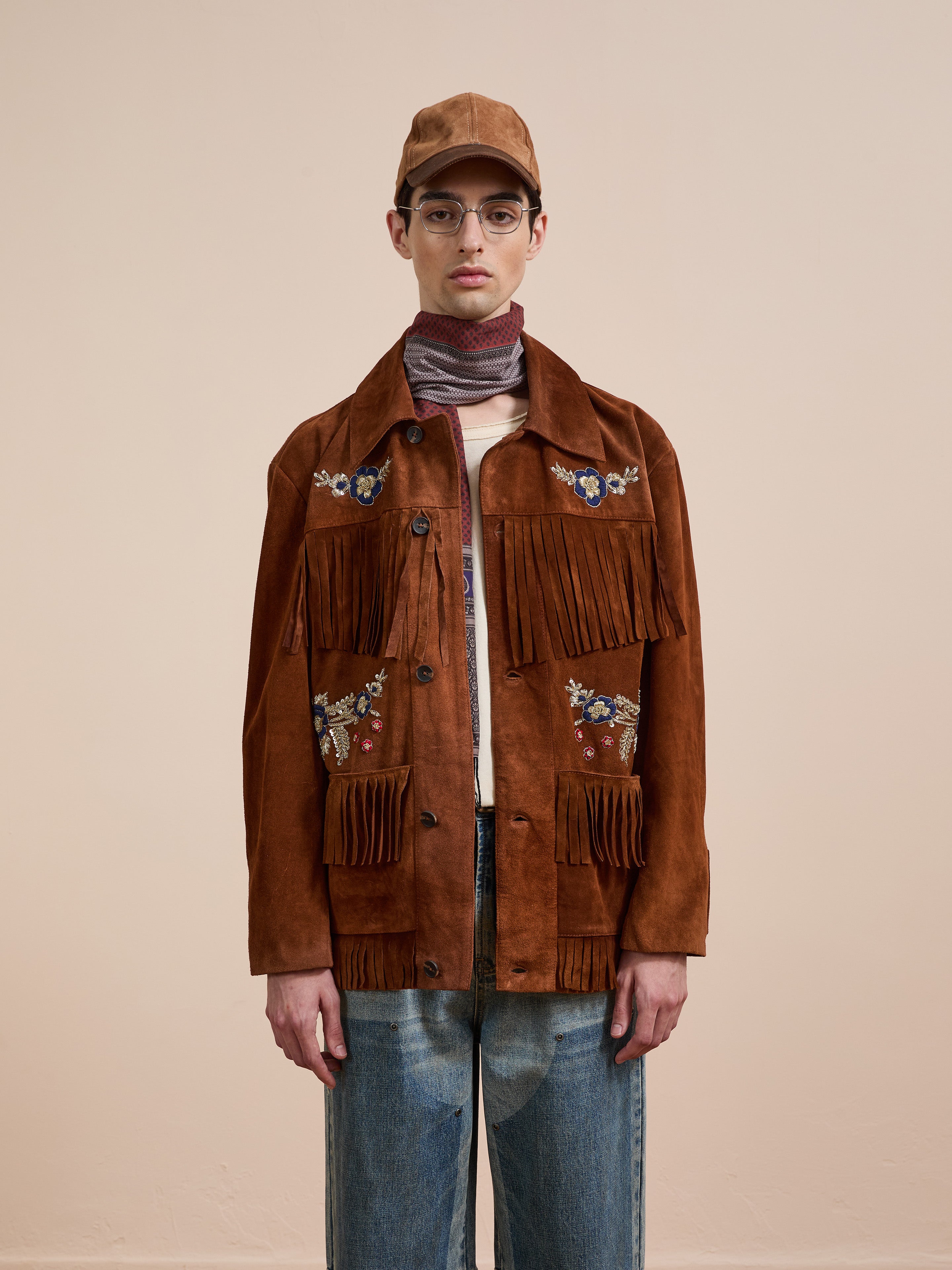 Wearing the FOUND Hand Embroidered Suede Fringe Jacket, blue jeans, a brown cap, glasses, and a striped scarf, a person stands against a plain beige background.