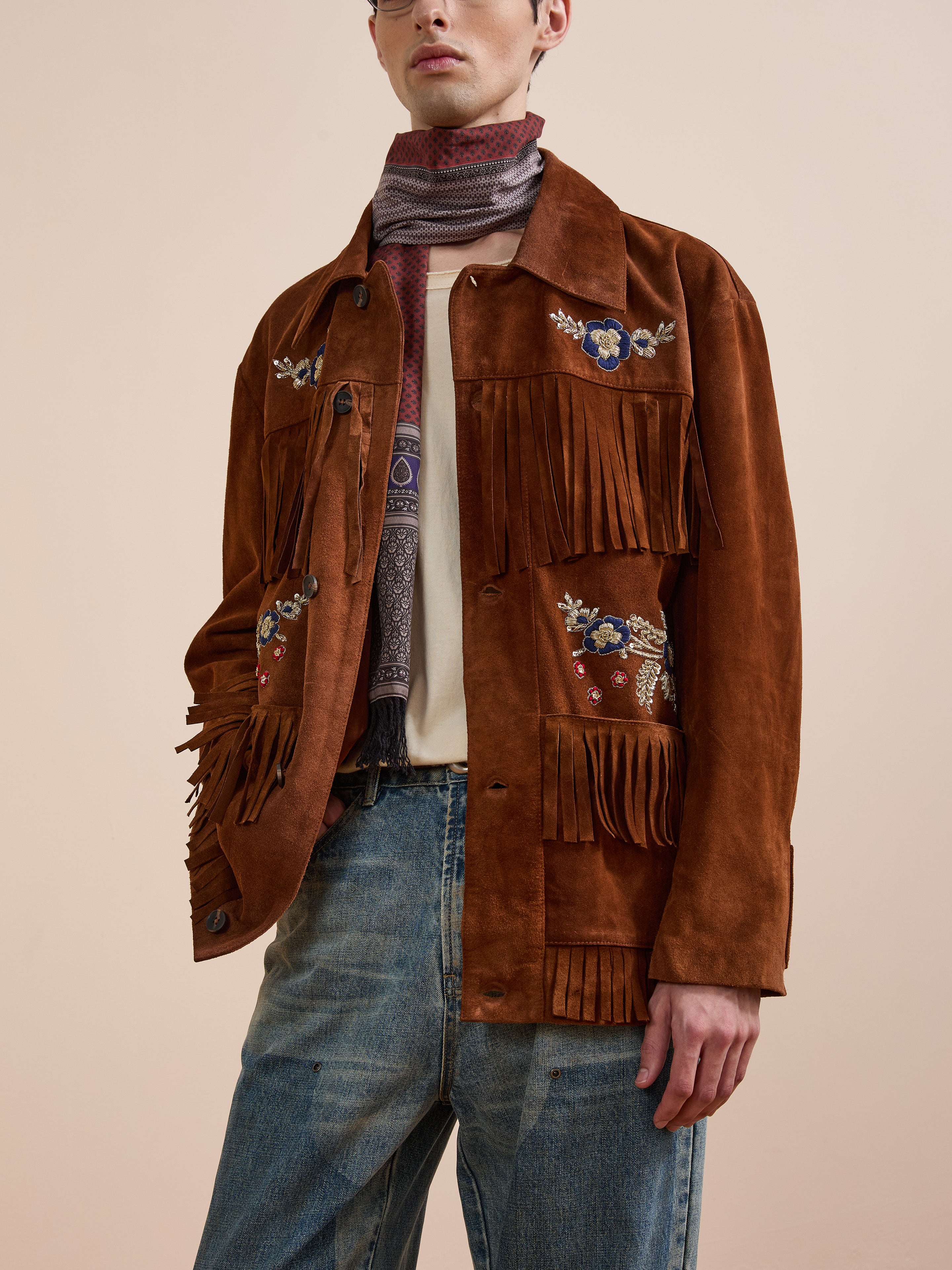 A person wearing the FOUND Hand Embroidered Suede Fringe Jacket with blue jeans, a light shirt, and a striped scarf stands against a plain background.