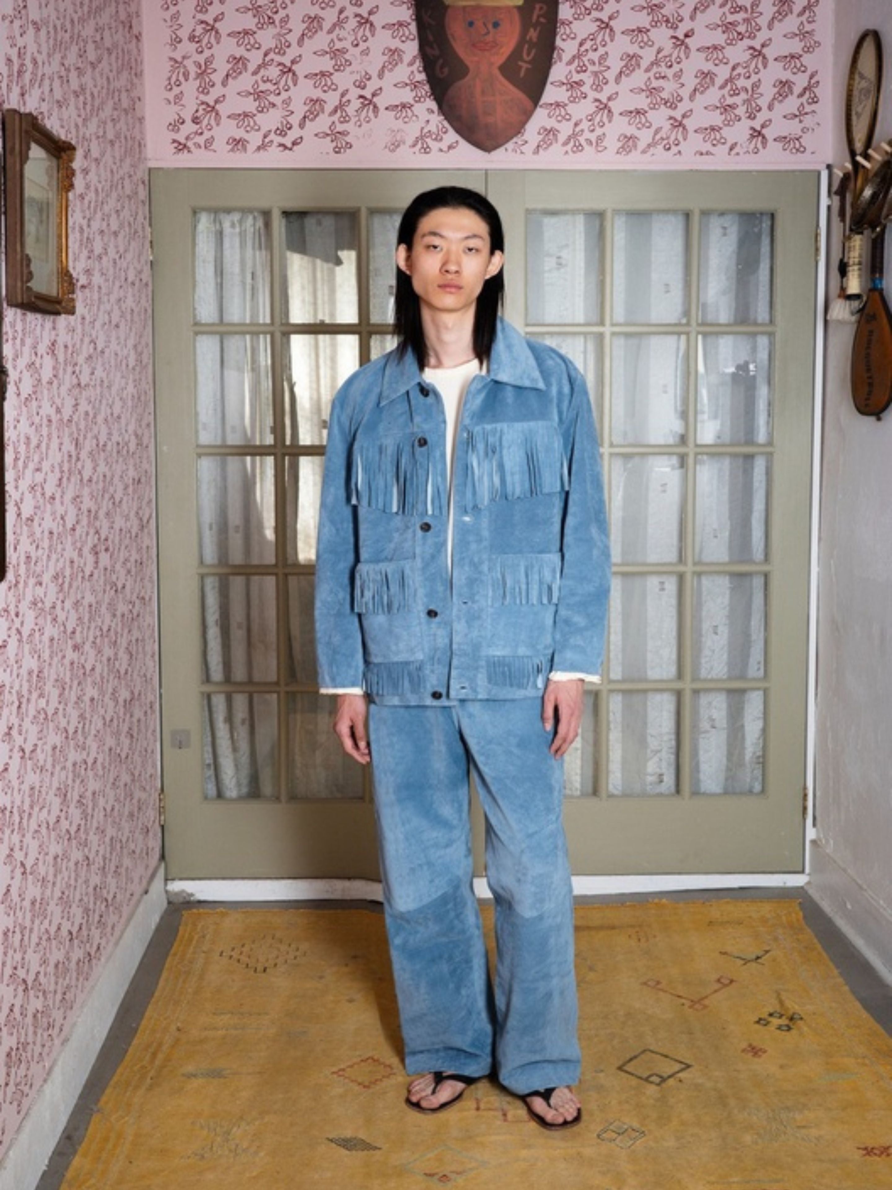 A person stands indoors on a yellow rug, wearing FOUND’s Western Suede Fringe Jacket in light blue with matching pants, white shirt, and flip-flops, set against floral wallpaper and glass doors.