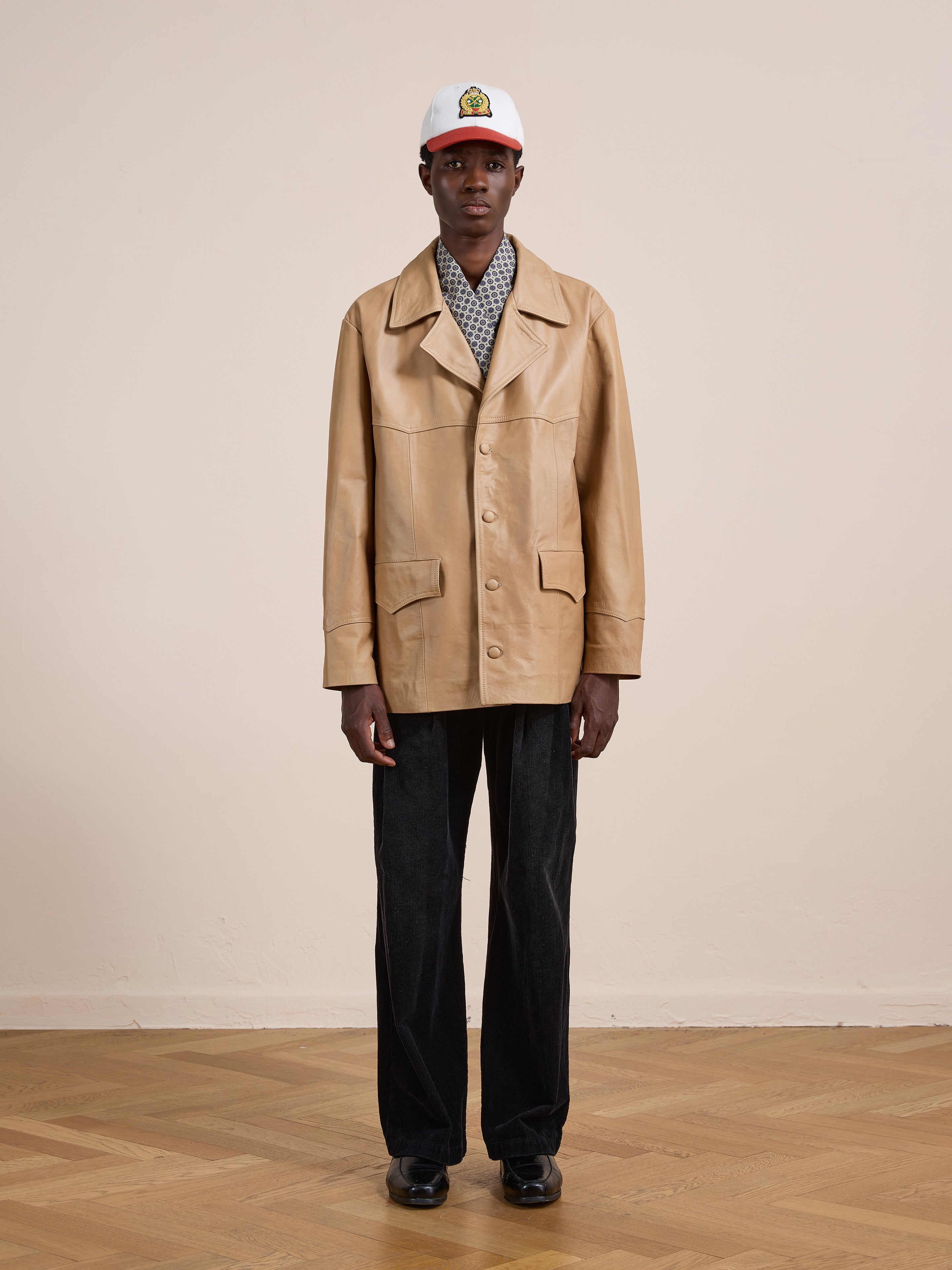 Indoors, a person wears the FOUND Western Oversized Leather Blazer in tan, styled with a patterned shirt, black pants and shoes, plus a white baseball cap featuring a colorful logo.