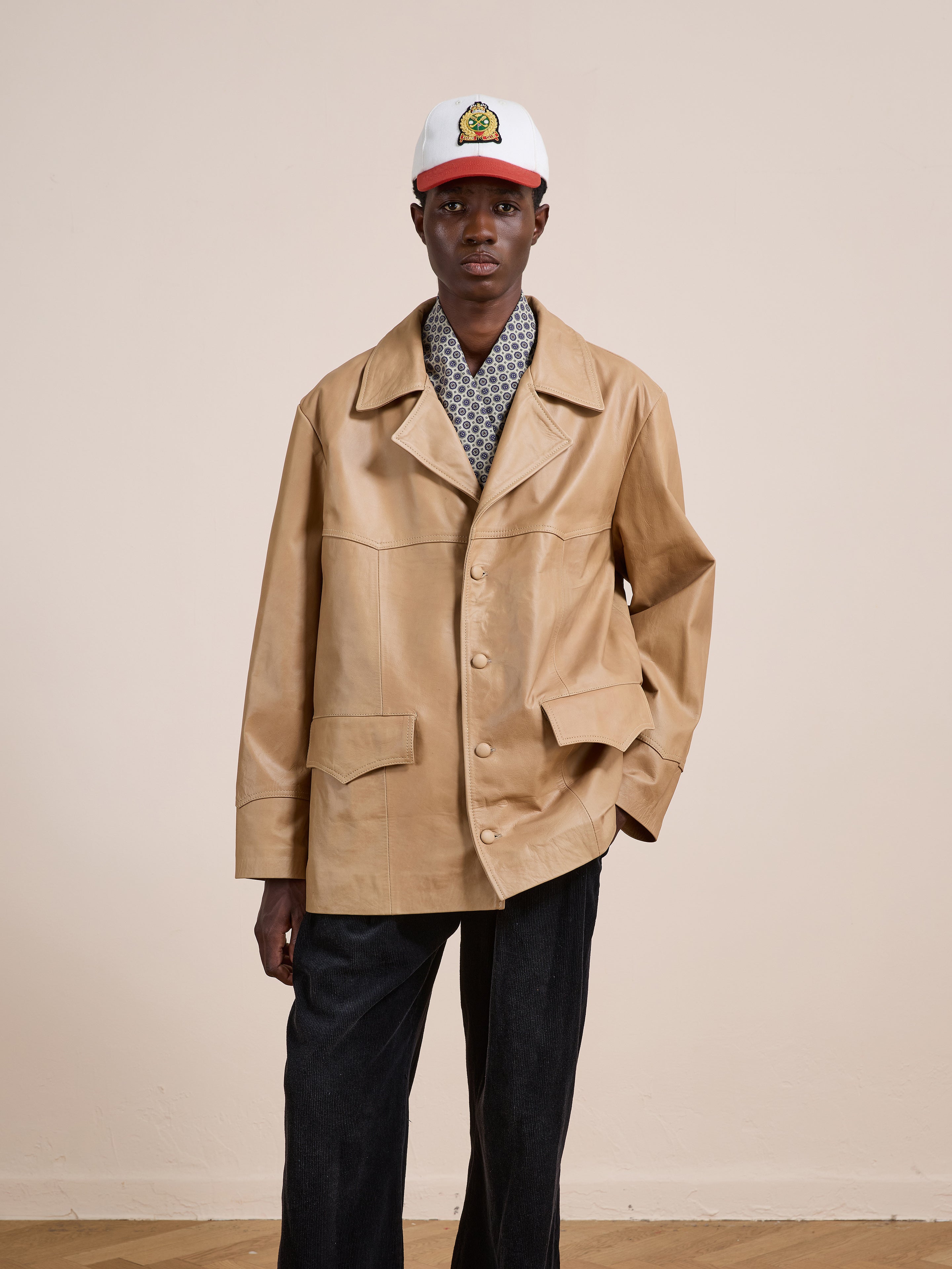 A person wears the FOUND Western Oversized Leather Blazer in beige over a patterned shirt, paired with dark pants and a white cap, standing against a plain light-colored background.