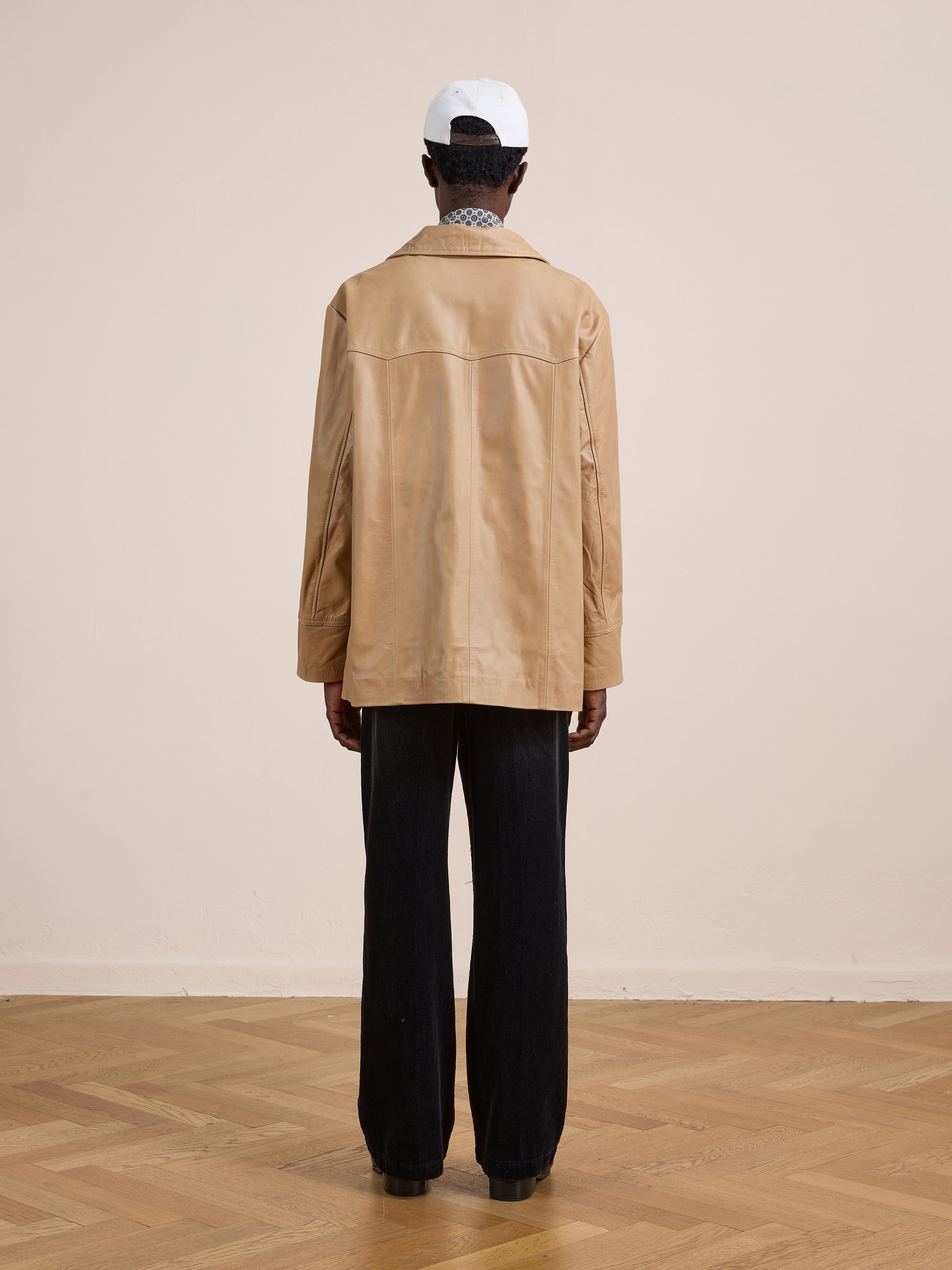 A person faces away, wearing the FOUND Western Oversized Leather Blazer in tan, paired with black pants and a white baseball cap, standing on a wooden floor before a plain beige wall.