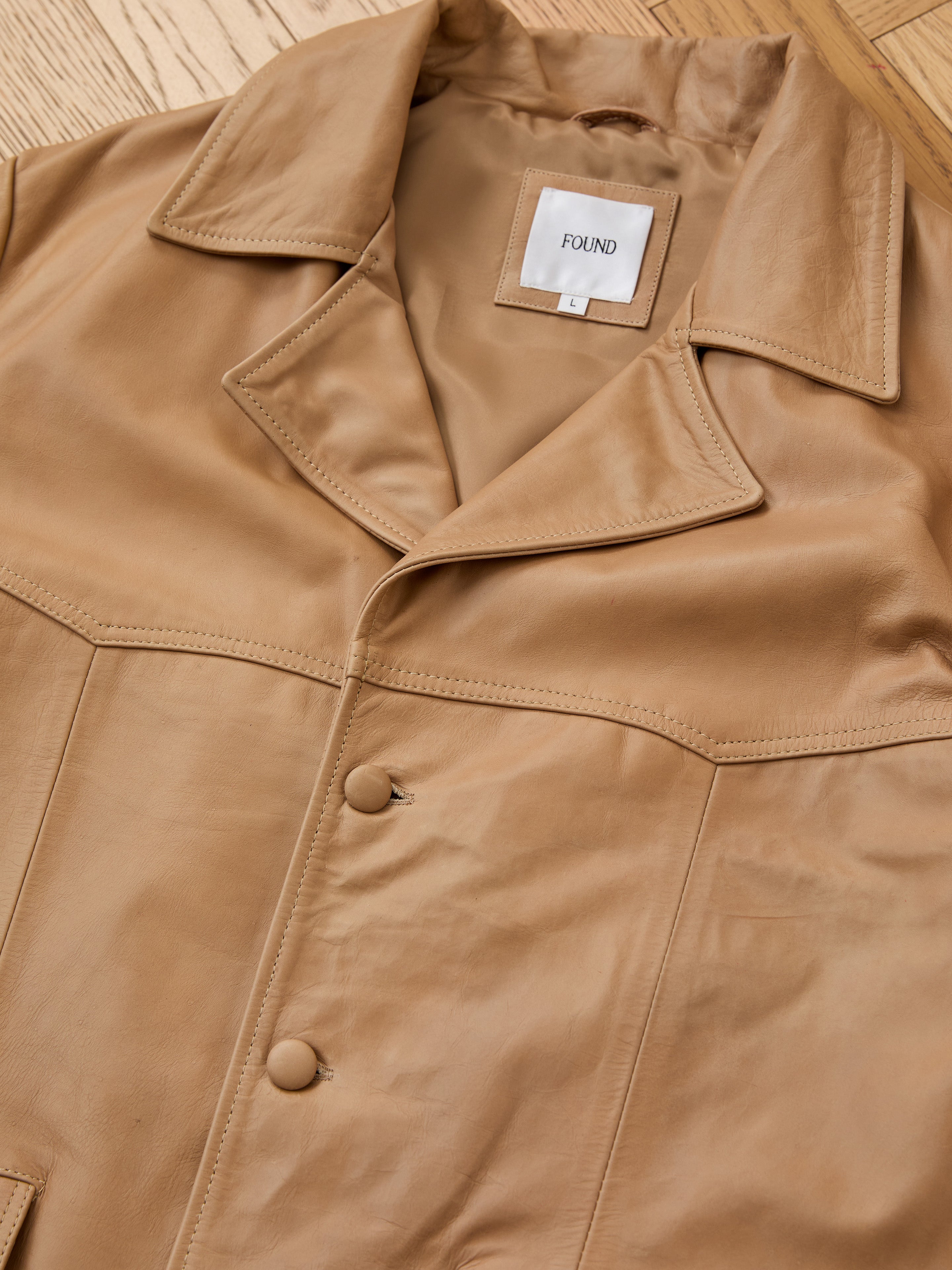 The FOUND Western Oversized Leather Blazer, in tan leather with large lapels and two buttons, is displayed flat on a wooden surface. Its label reads "FOUND.
