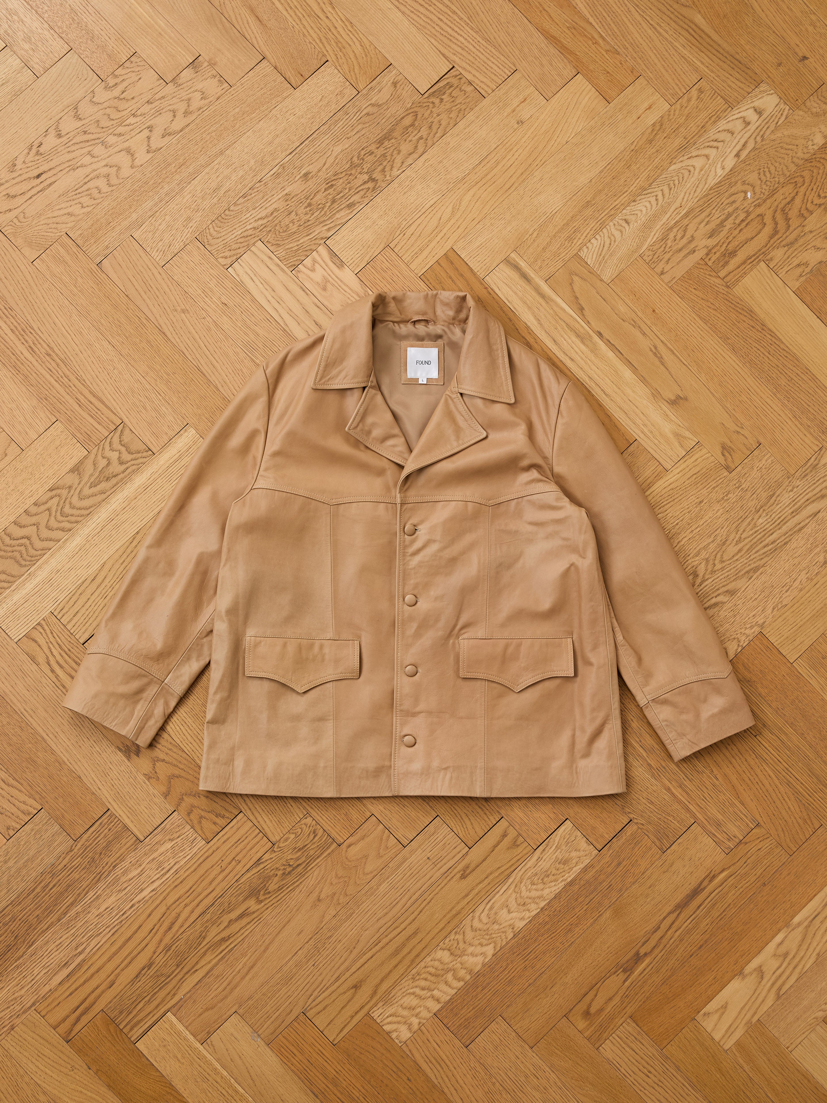The FOUND Western Oversized Leather Blazer, a tan button-up with a collar and two large front pockets, is laid flat on a wooden parquet floor.