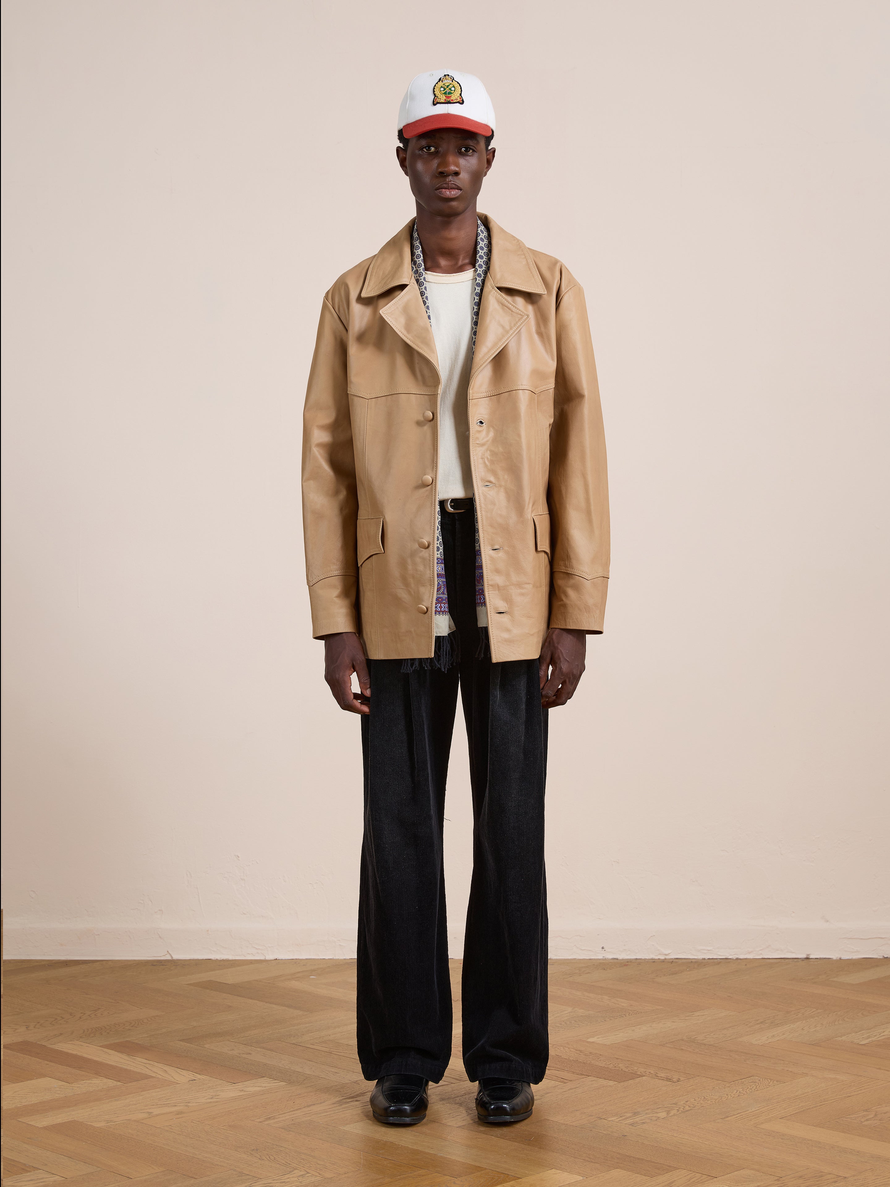 Indoors against a plain light wall, a person wears the FOUND Western Oversized Leather Blazer in tan, styled with a white T-shirt, dark wide-leg pants, black shoes, and a white patch baseball cap.