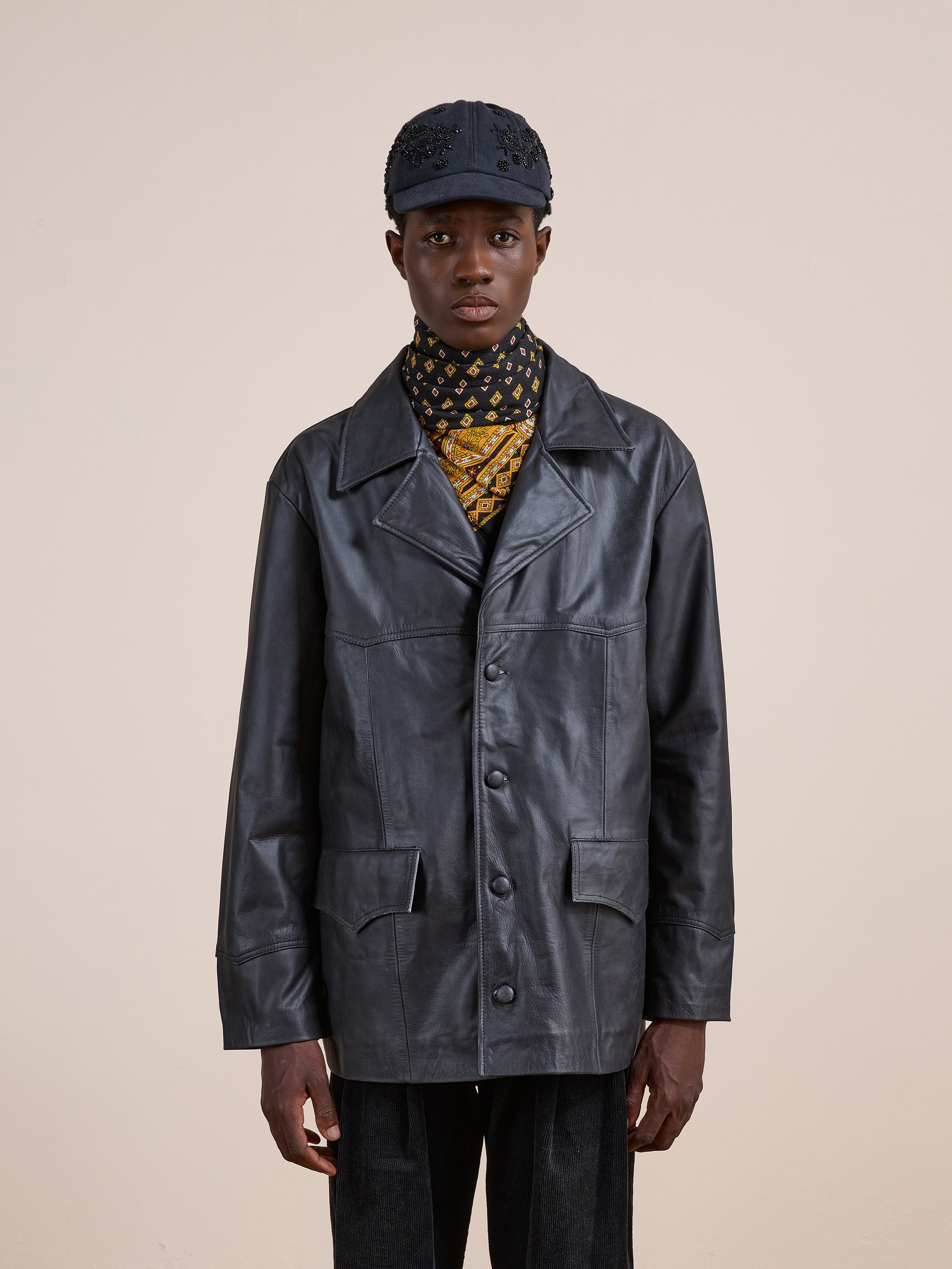 A person wears the FOUND Western Oversized Leather Blazer styled with black pants, a black cap, and a patterned yellow and black scarf, standing against a plain beige background.