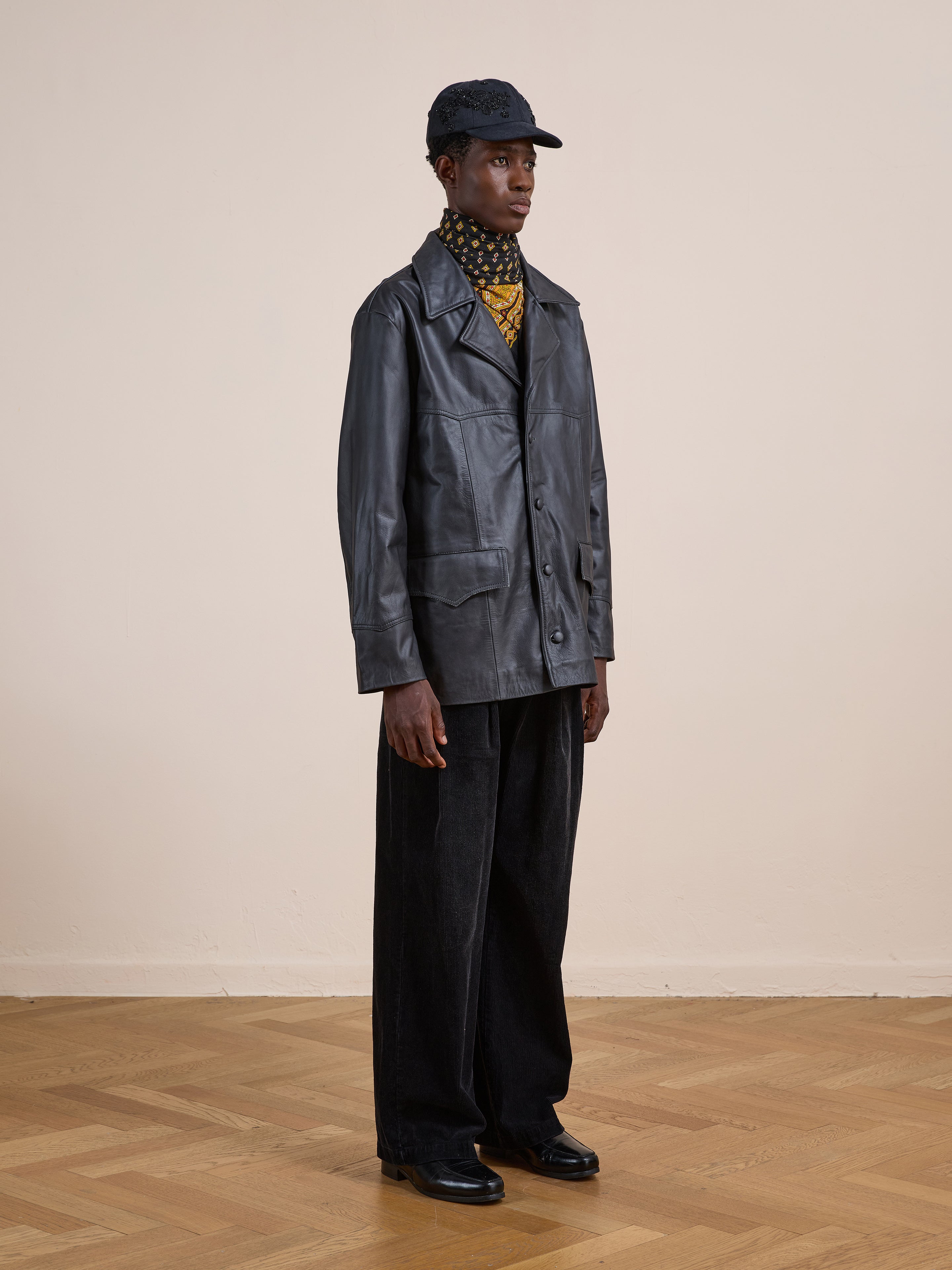Indoors against a beige wall, a person wears FOUND’s Western Oversized Leather Blazer with wide-leg dark pants, black shoes, a patterned turtleneck, and a black cap.