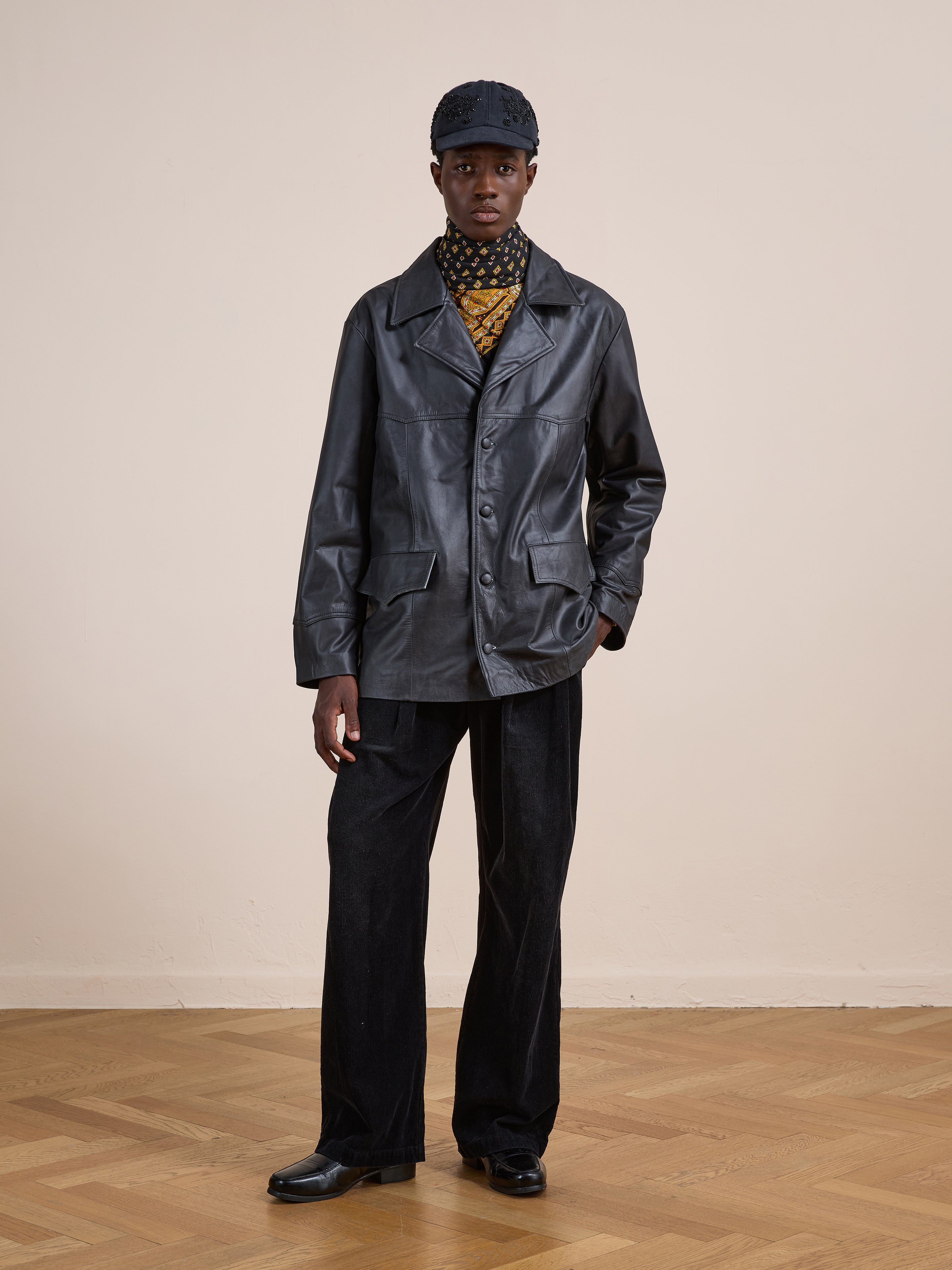 A person stands indoors on a wooden floor against a beige wall, wearing the FOUND Western Oversized Leather Blazer, styled with black pants, a patterned scarf, black cap, and black shoes.