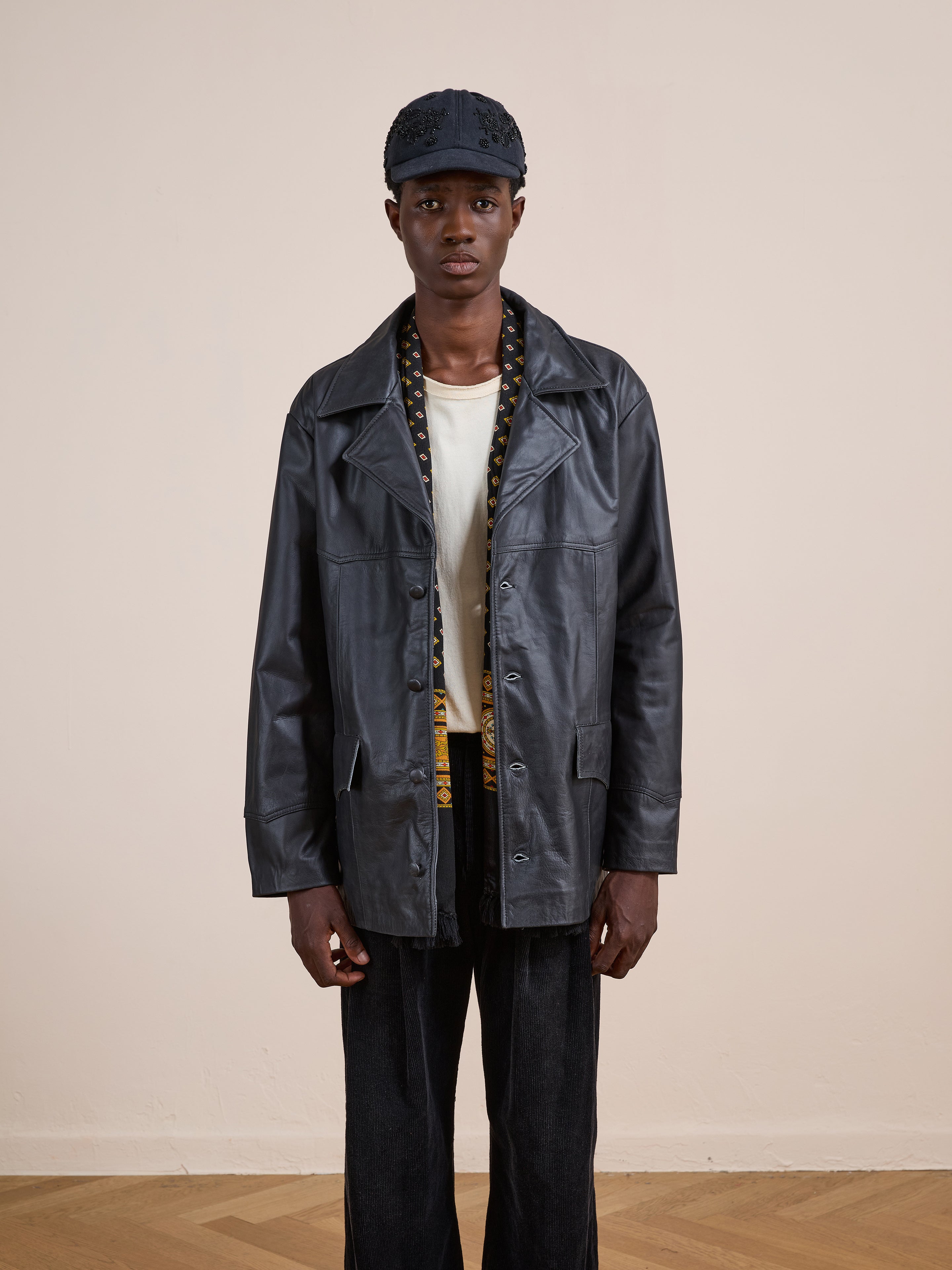 A person in the FOUND Western Oversized Leather Blazer, white shirt, patterned scarf, black pants, and a black cap stands against a plain, light-colored wall.