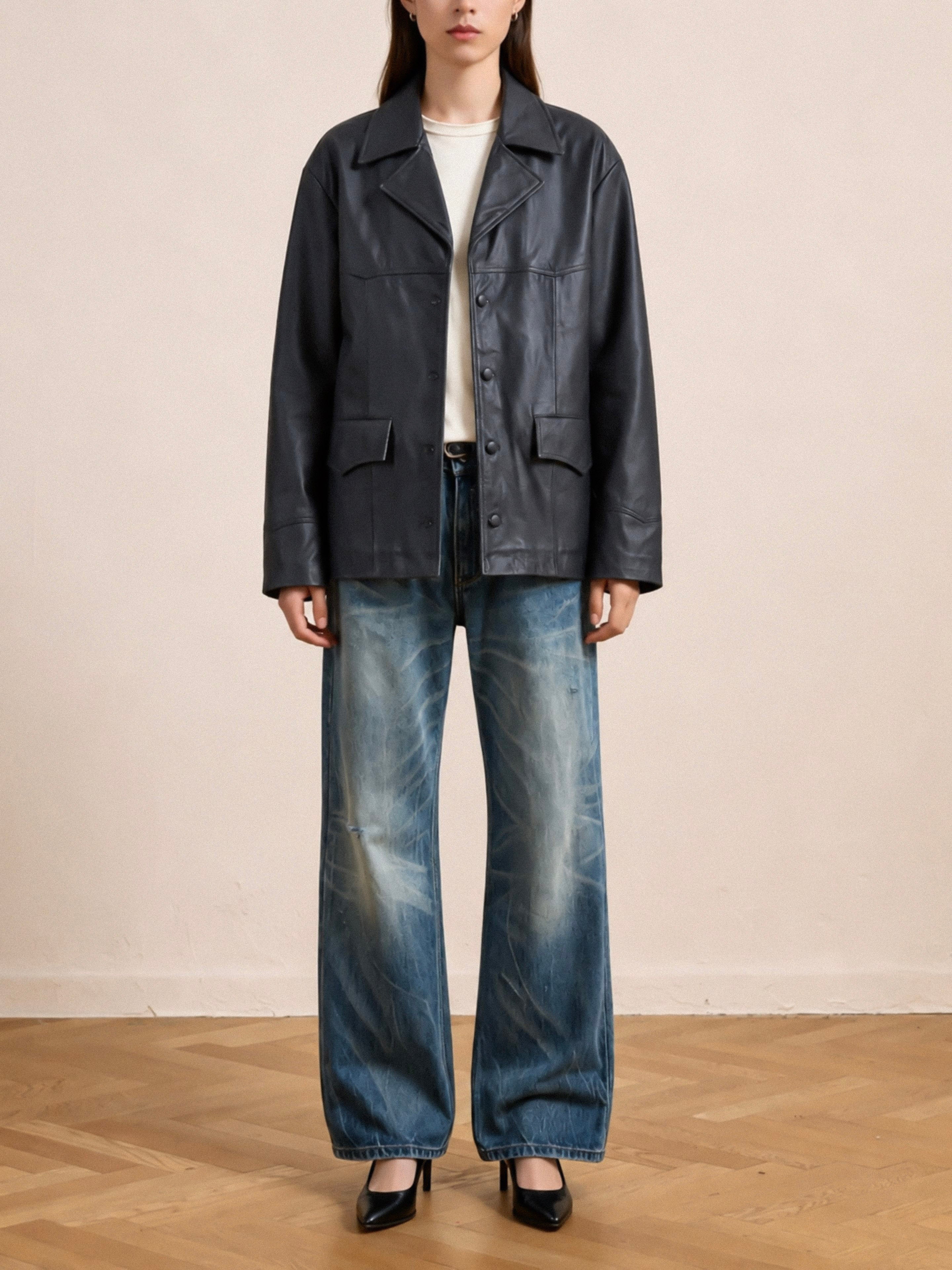 A person stands indoors against a plain beige wall and wooden floor, wearing the FOUND Western Oversized Leather Blazer in black styled over a white shirt, paired with wide-leg faded blue jeans and black heels.