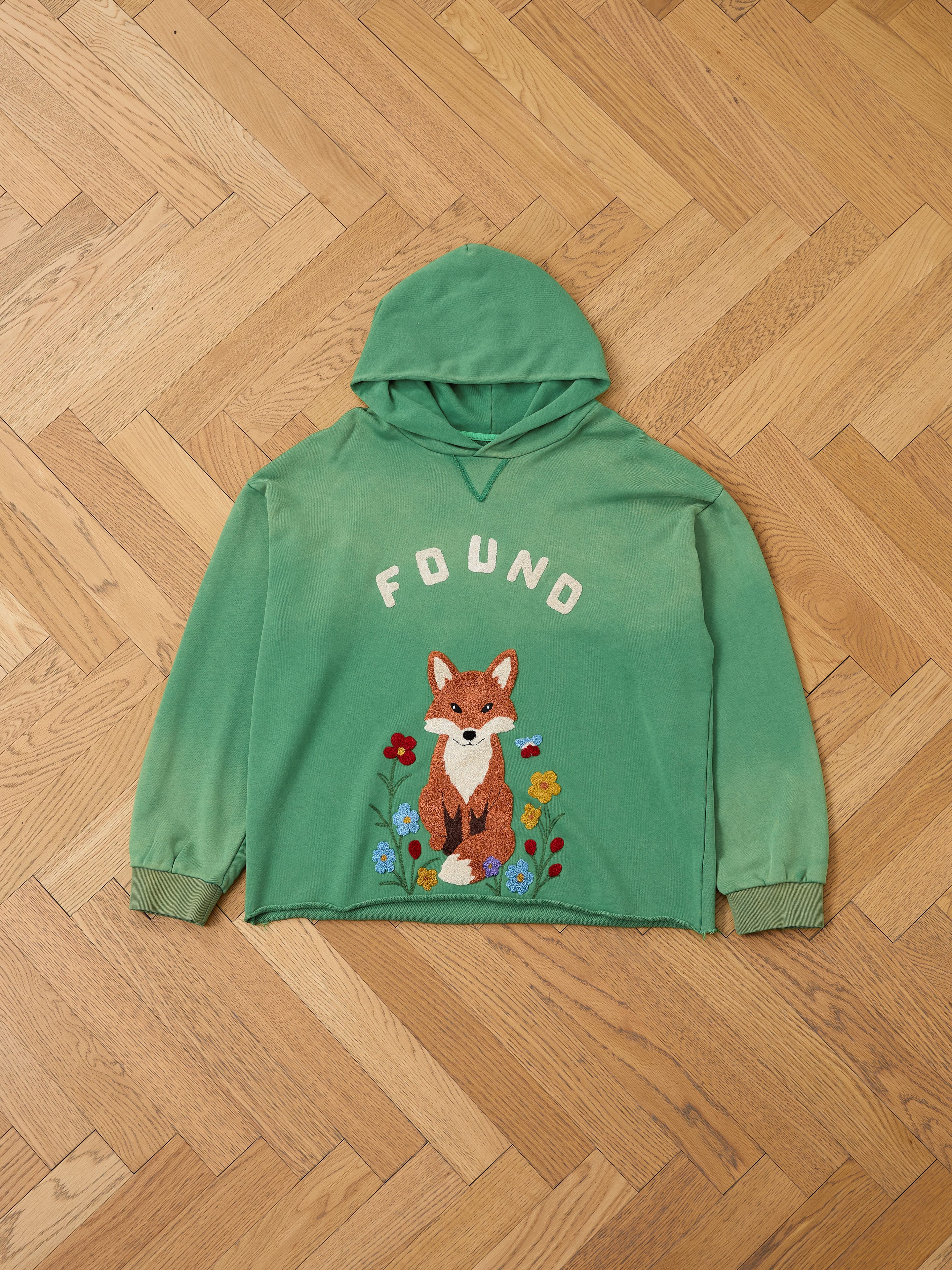 The FOUND Embroidered Wildlife Hoodie features a green unisex relaxed fit in soft French-terry cotton, "FOUND" text, and an embroidered fox with flowers, shown here on a wooden parquet floor.