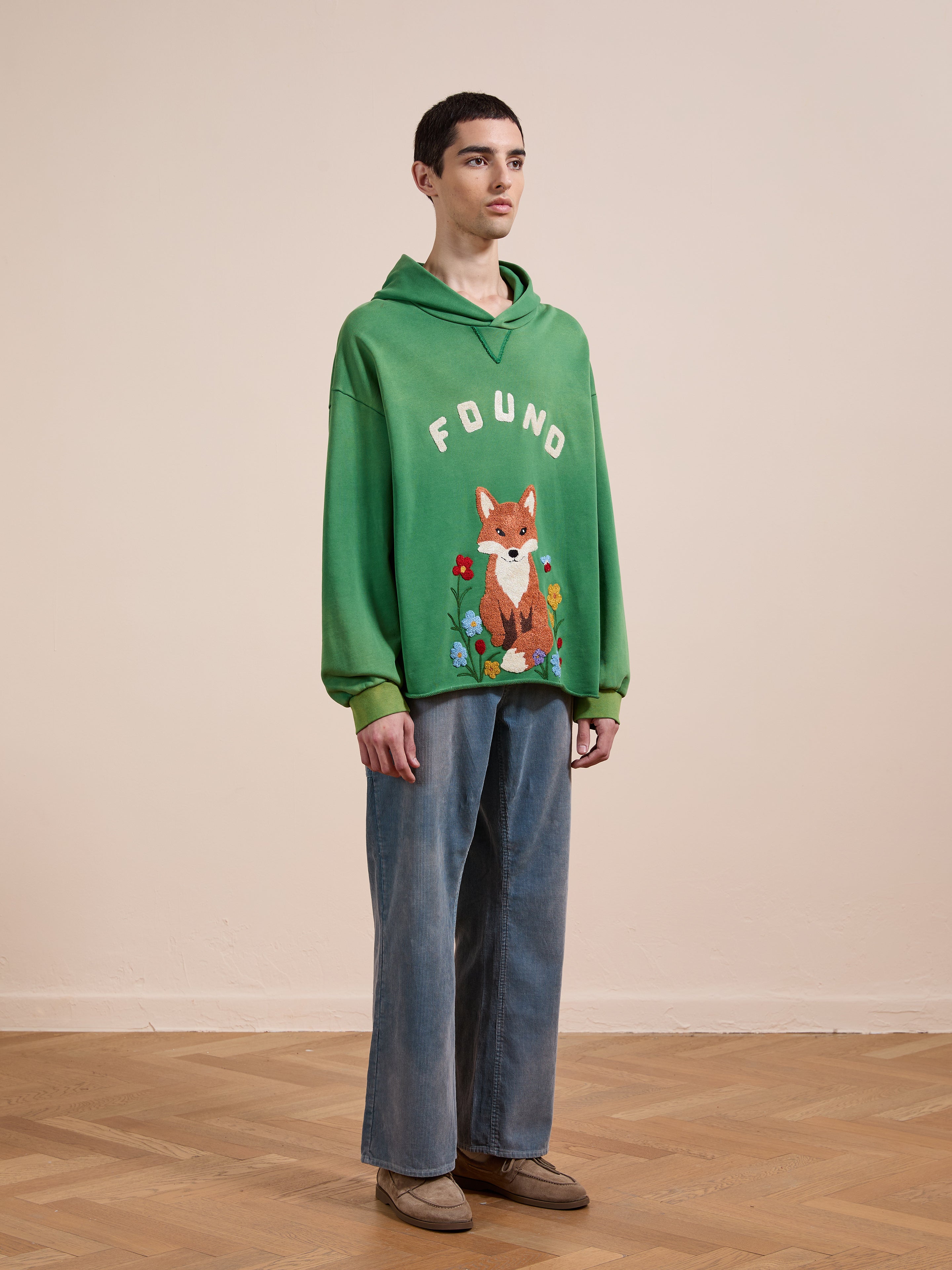 Indoors against a beige wall, a person wears the FOUND Embroidered Wildlife Hoodie in green with a fox graphic, paired with blue wide-leg jeans and brown shoes.