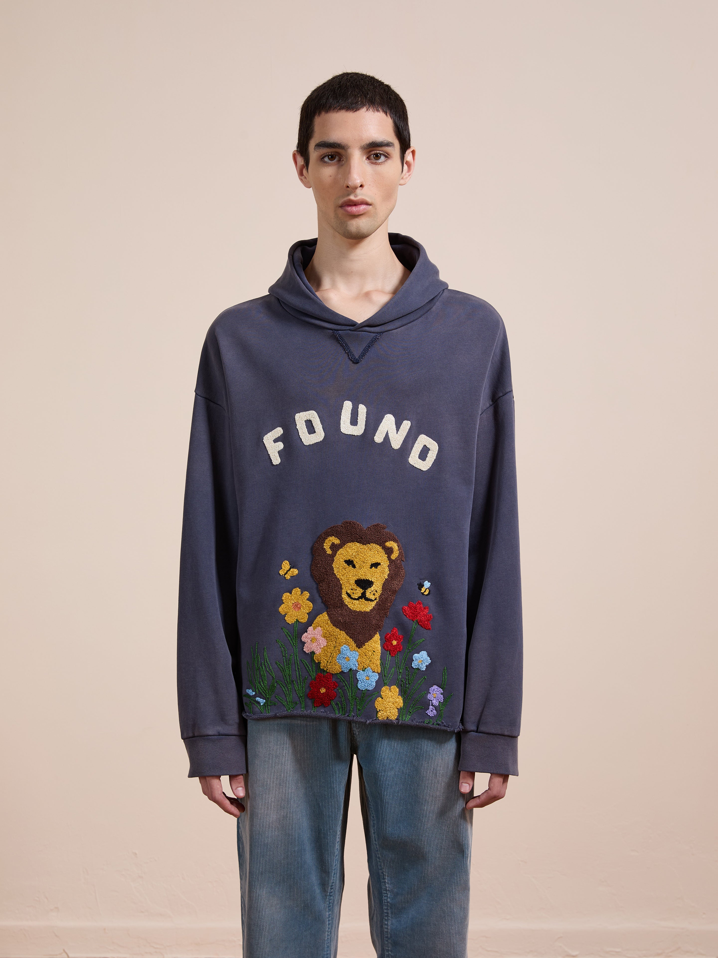 A person stands against a plain background wearing the FOUND Embroidered Wildlife Hoodie, featuring a lion with flowers and "FOUND" text on Vintage French-terry Cotton, paired with blue jeans.