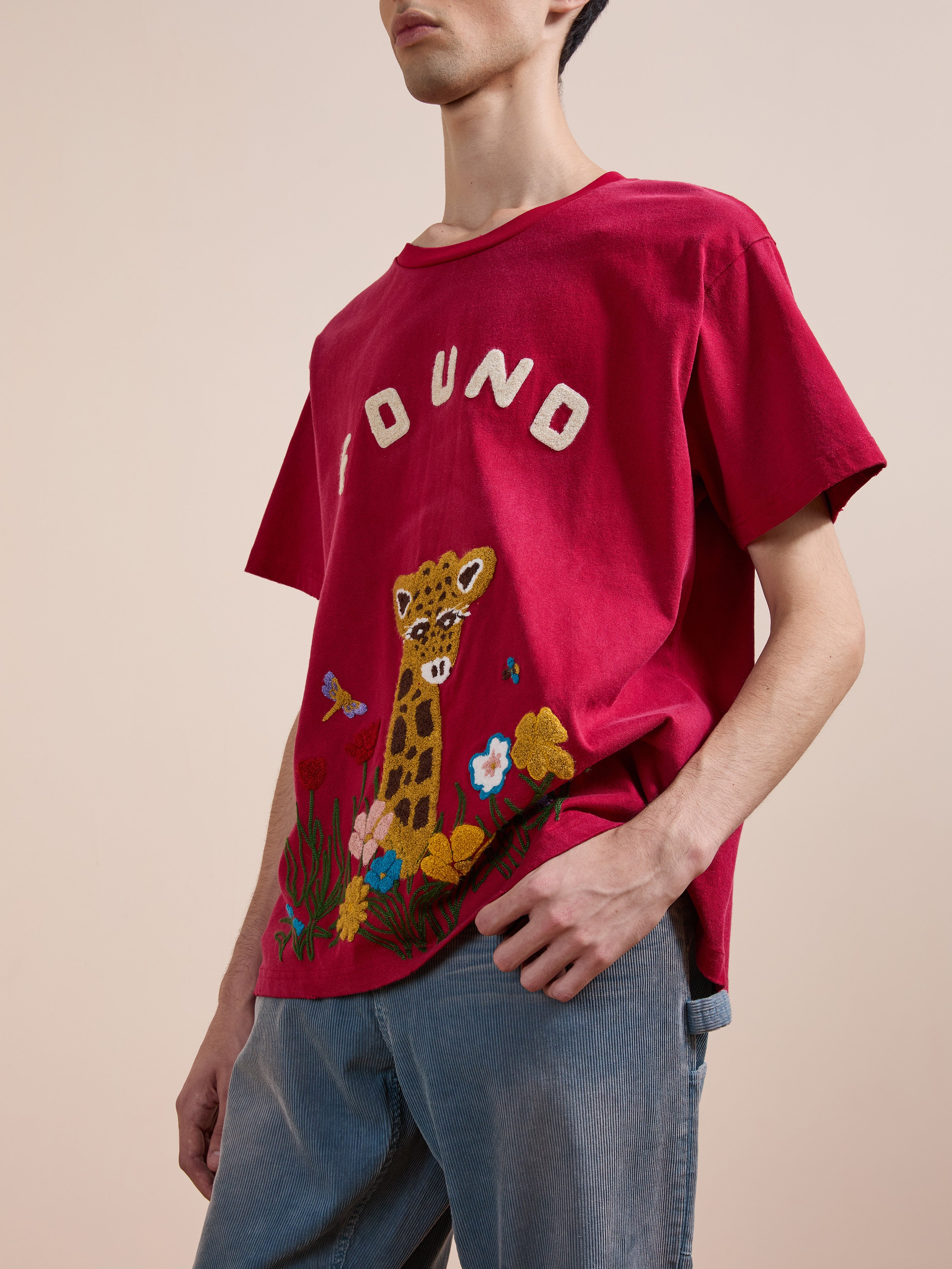 Someone wearing a red FOUND Embroidered Wildlife Tee featuring a giraffe, flowers, and butterflies graphic, paired with light blue jeans.