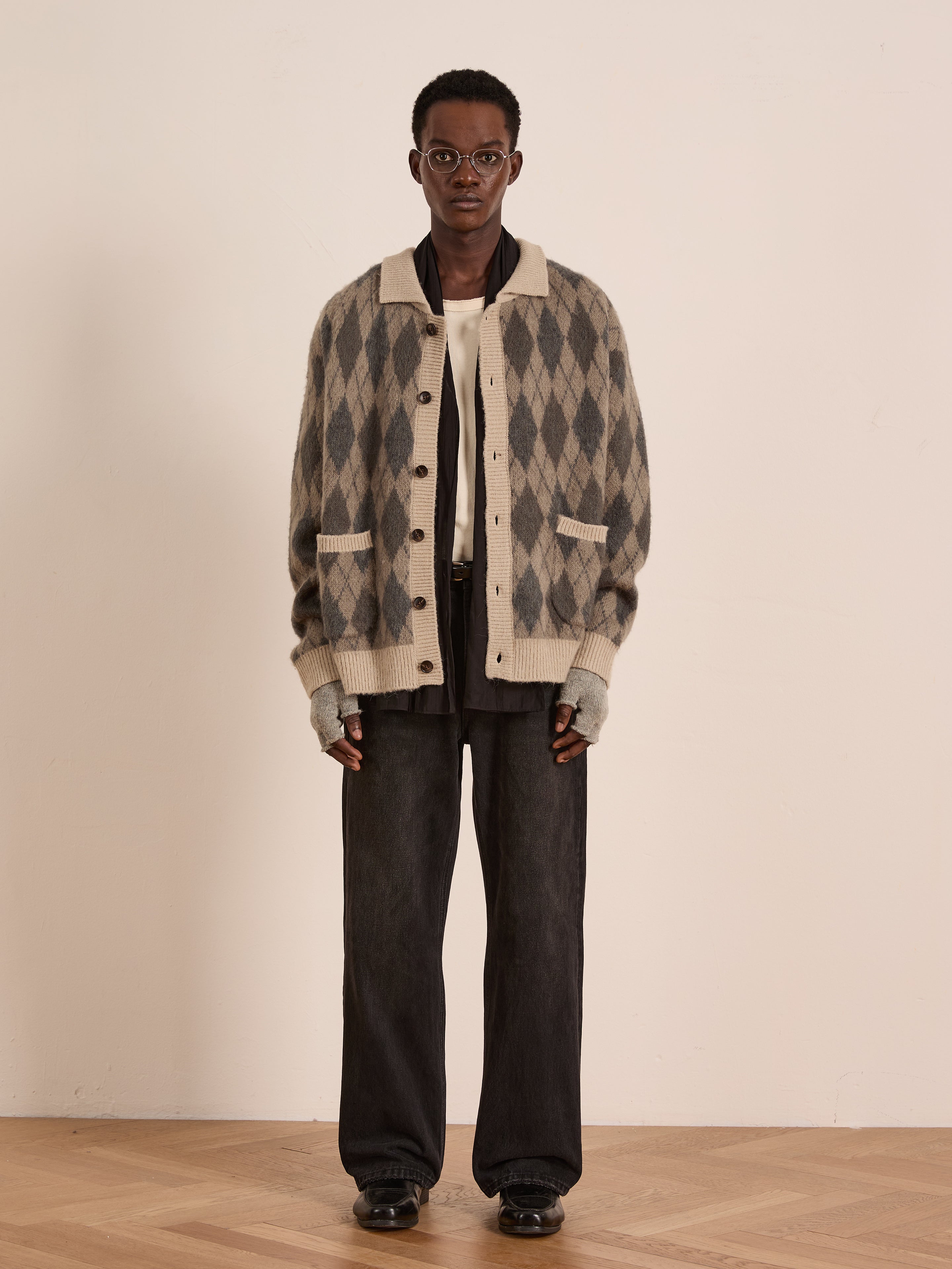 Indoors against a plain light background, a person wears glasses, wide-leg dark pants, fingerless gloves, black shoes, a white shirt, and the FOUND Argyle Cardigan.