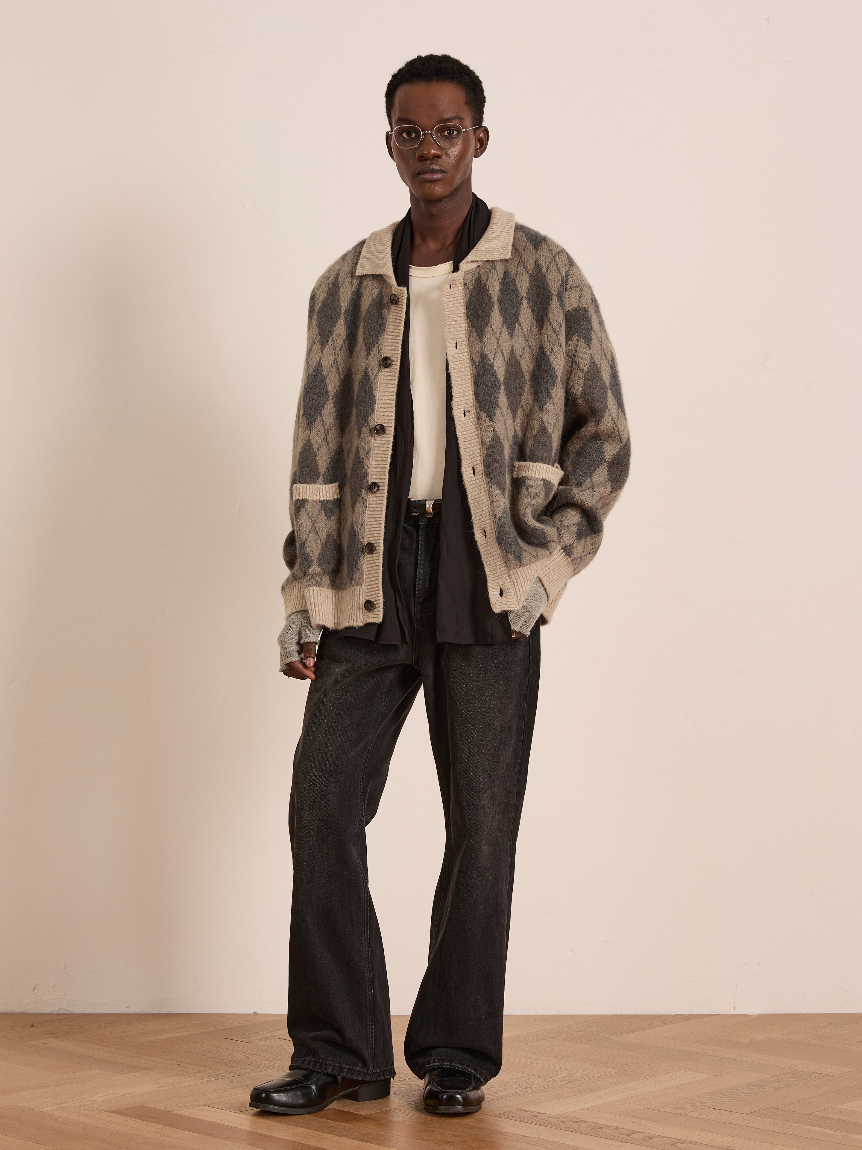 A person wearing glasses stands against a light wall, dressed in the FOUND Argyle Cardigan—a relaxed fit in beige and gray wool blend—layered over a white shirt, with black jeans and black shoes.