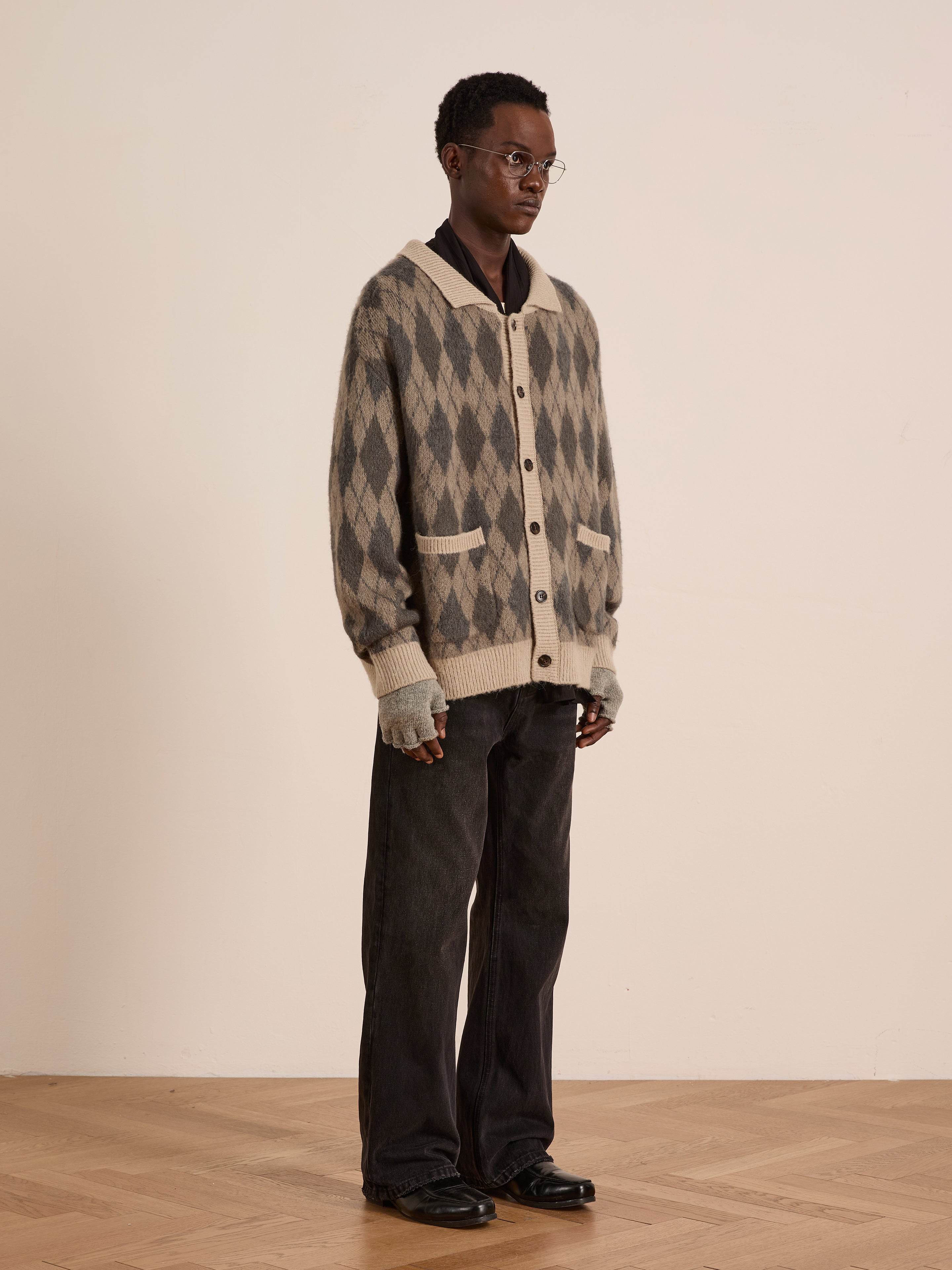 Indoors, a person wearing glasses pairs fingerless gloves, black shirt, loose black jeans, and black shoes with the FOUND Argyle Cardigan against a plain light background.