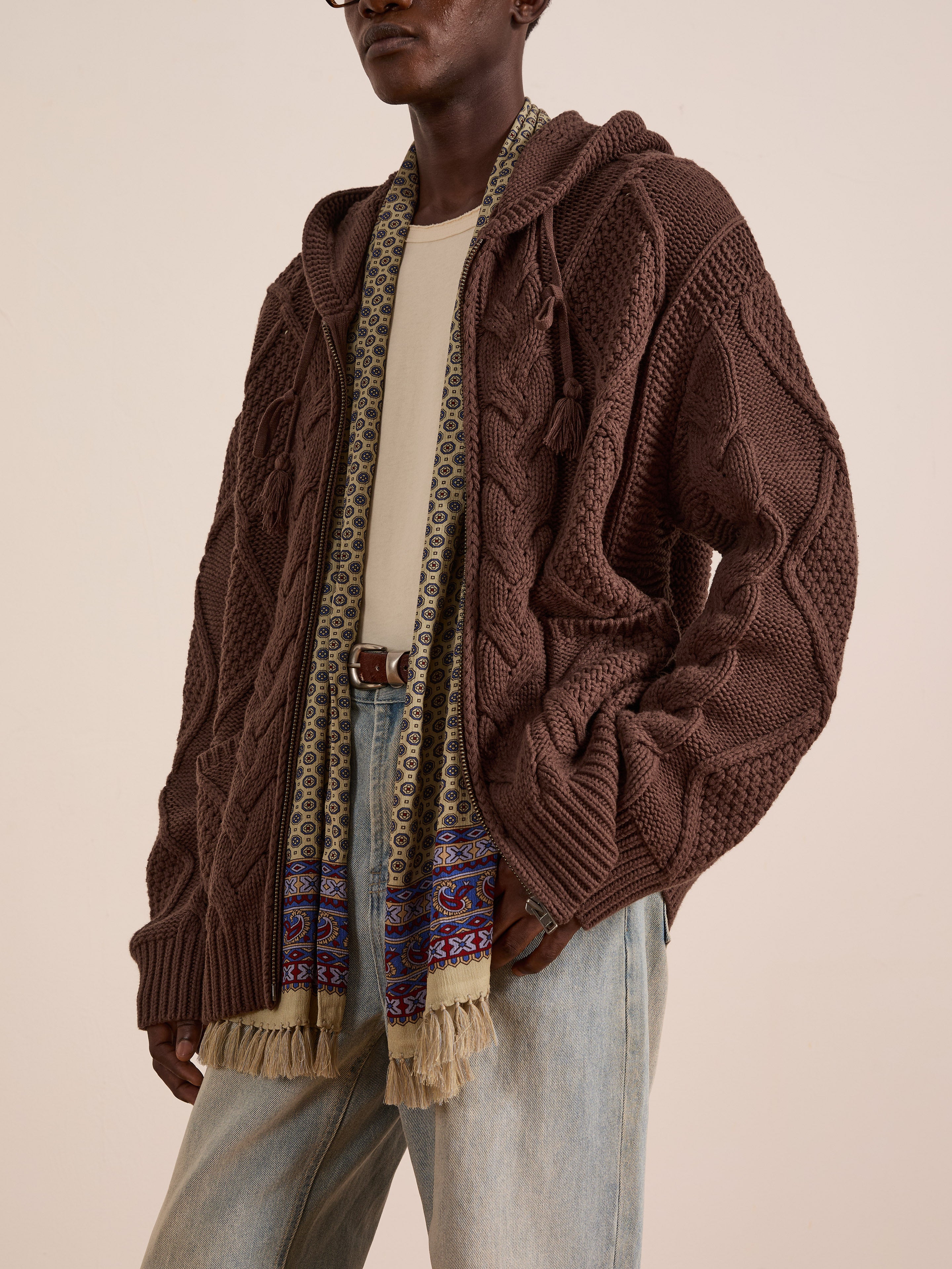 Wearing the FOUND Zip Up Cable Knit Hoodie—a brown, vintage-inspired, chunky unisex piece with a relaxed fit—layered over a beige shirt, blue jeans, and a patterned fringe scarf.