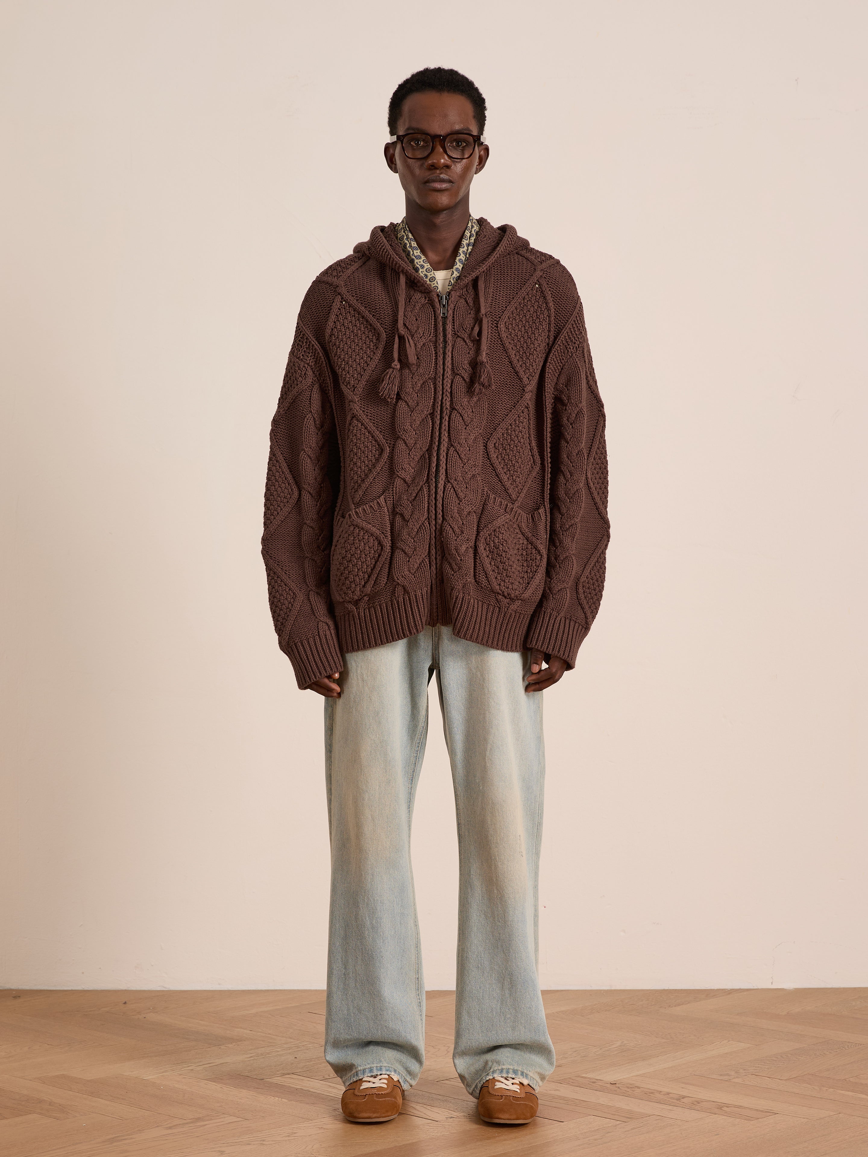 A person stands against a plain background wearing the FOUND Zip Up Cable Knit Hoodie, light blue wide-leg jeans, brown shoes, and glasses.