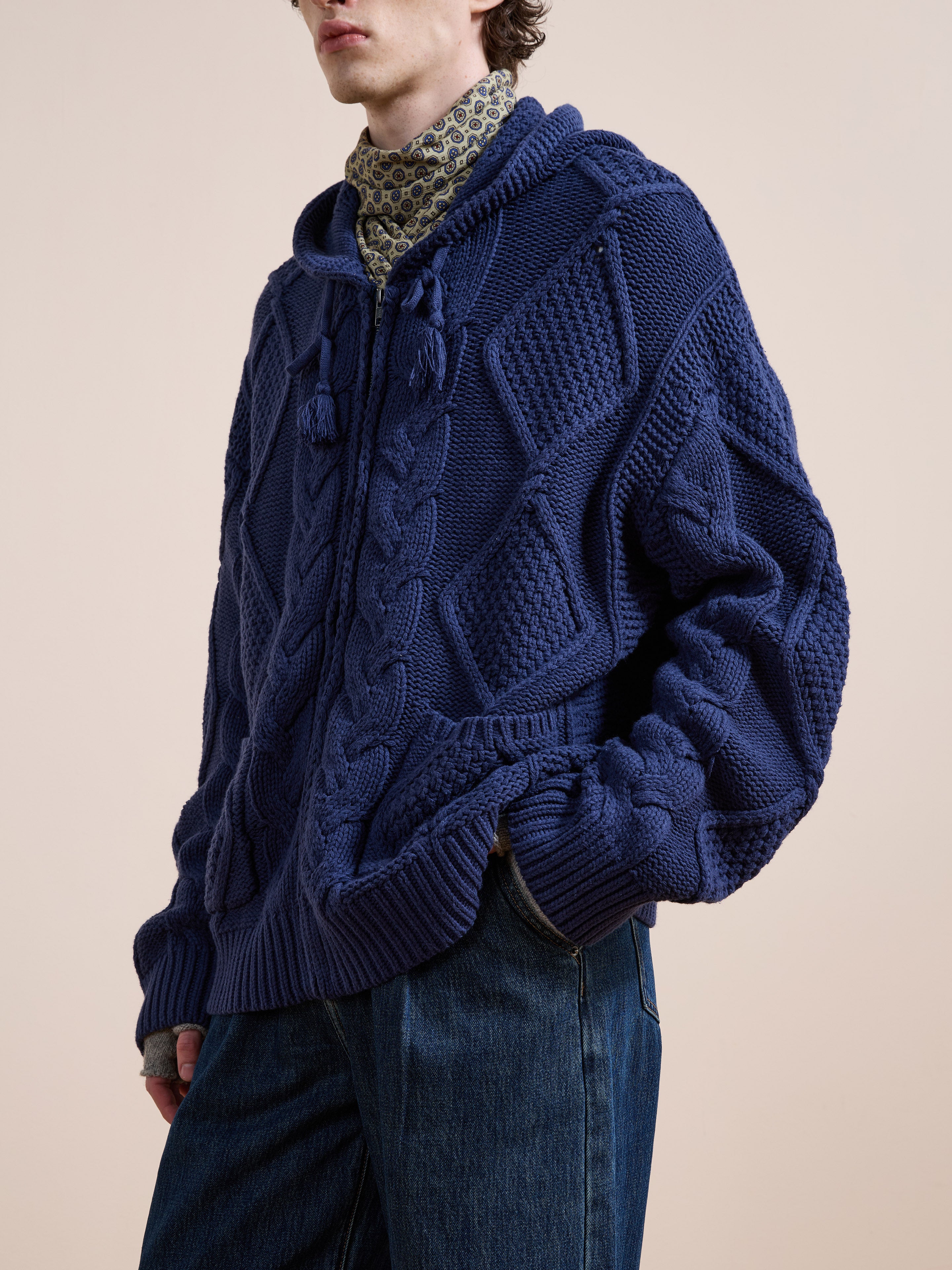 A person wearing the Found Zip Up Cable Knit Hoodie in textured blue, layered over a patterned high-neck shirt and blue jeans, stands against a plain background.