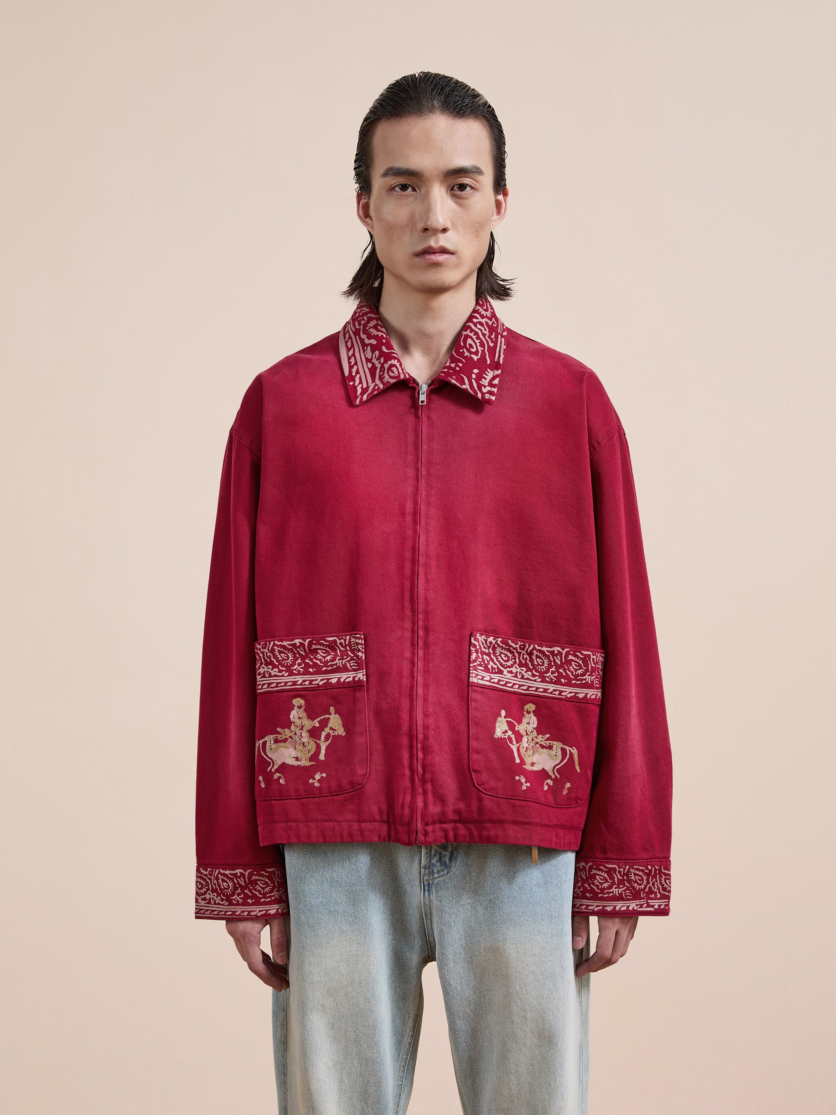 A person wears the Found Abhala Printed Work Jacket, featuring embroidered equestrian motifs and a unisex relaxed fit, paired with light blue jeans against a beige background.