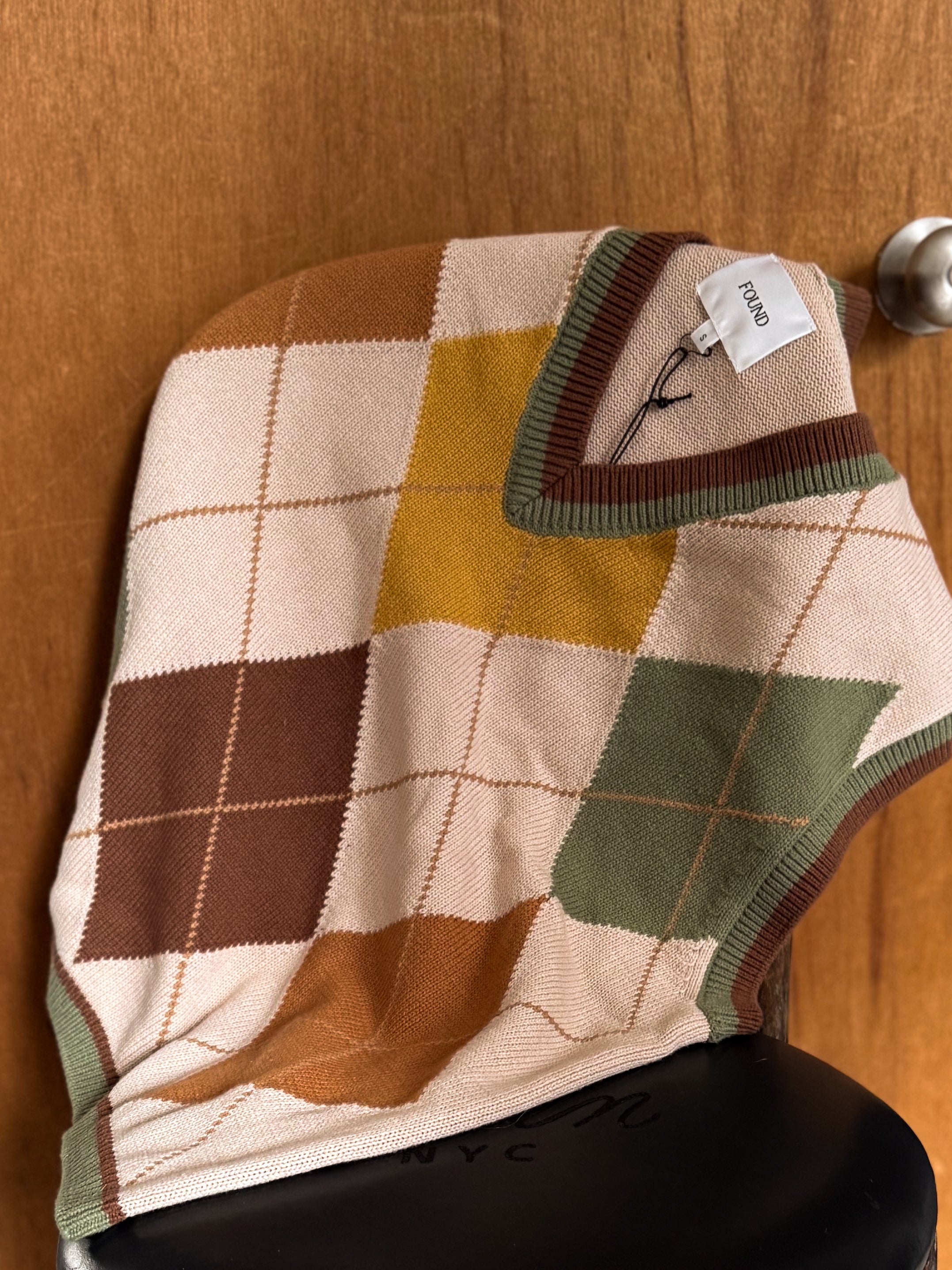 A FOUND Argyle Knit Vest with a vintage diamond pattern drapes over a black chair, exuding a collegiate vibe. A "Proud" tag adds academic charm to the scene.