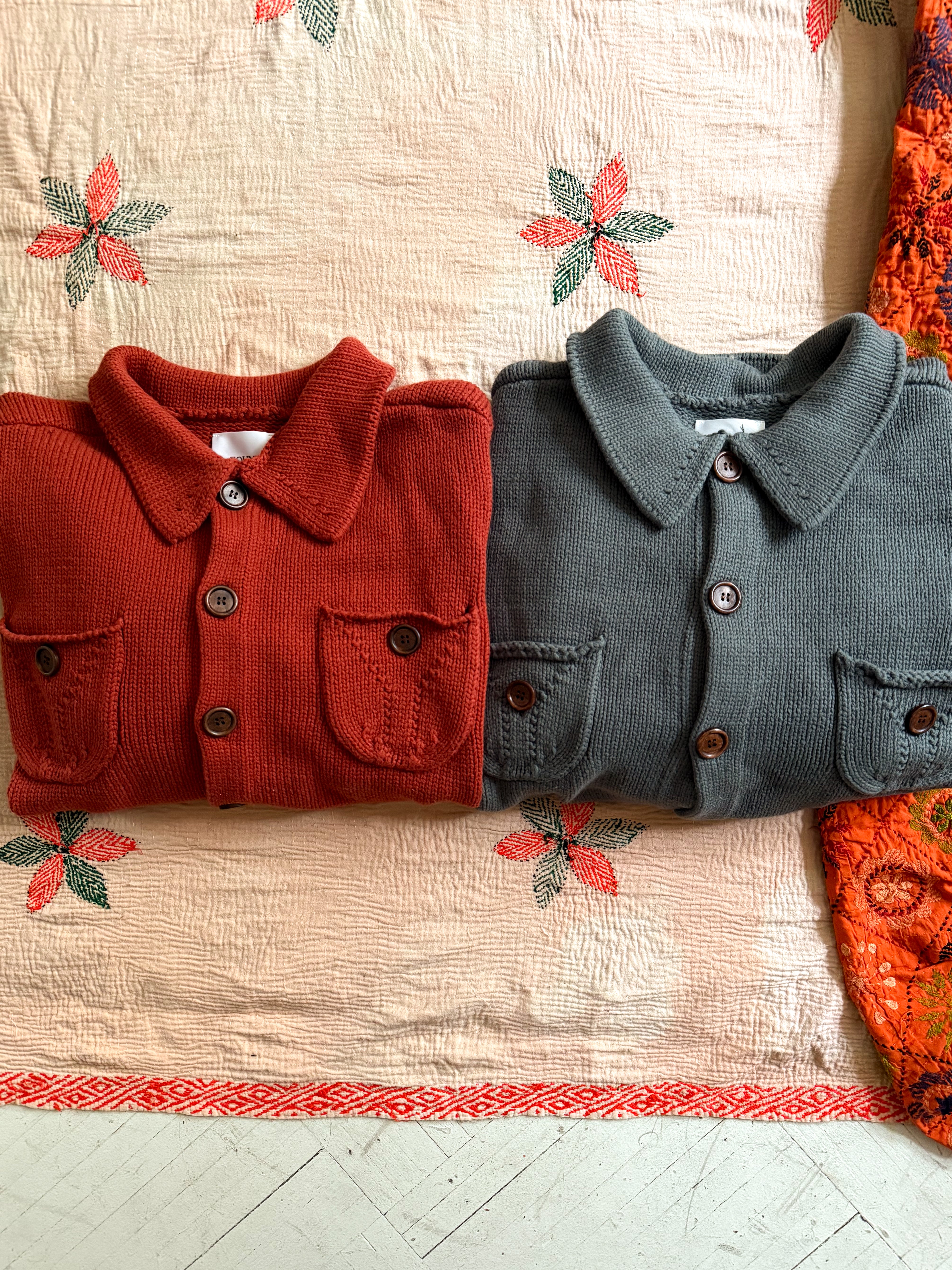 Two folded Hunter Knit Collared Cardigans by FOUND rest on a floral-patterned bedspread. The left is red and the right is gray, both mid-weight knits with buttoned pockets and collars, ideal for transitional layering.