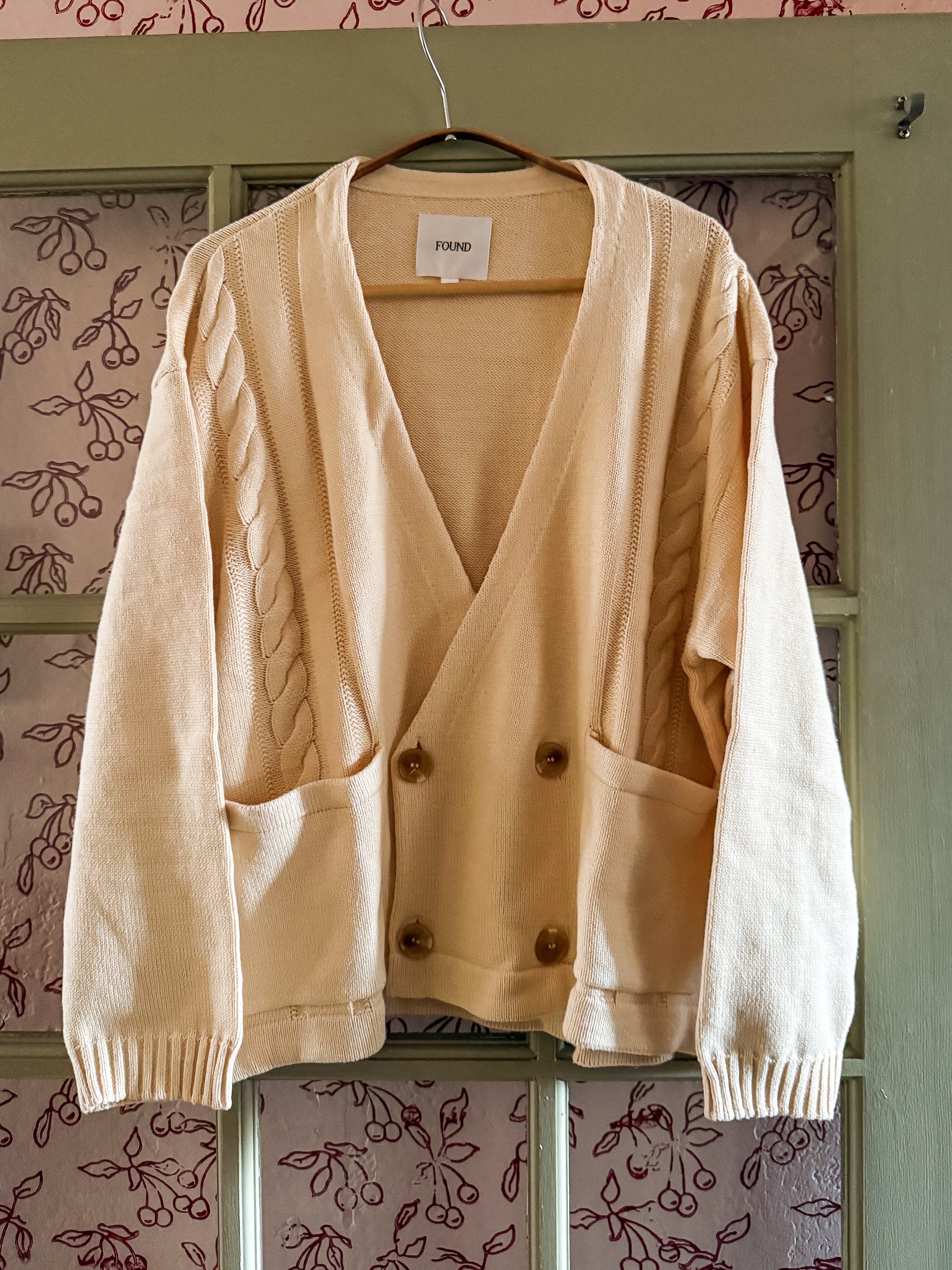 The FOUND Double Breasted Knit Cardigan in beige features cable knit detailing, a relaxed fit, and is made from a midweight cotton blend. It hangs on a hanger against a decorative backdrop with buttons down the front.