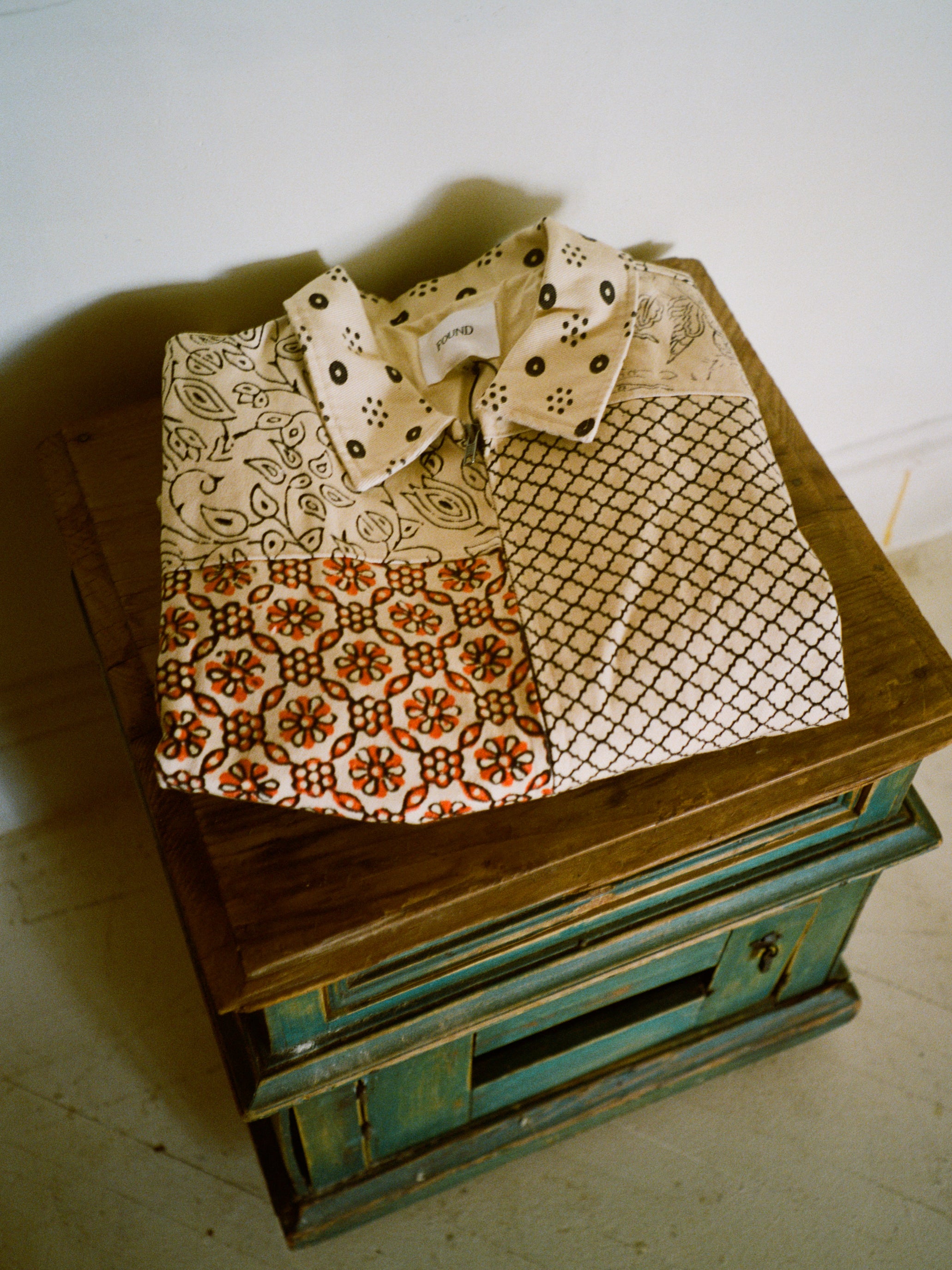 A folded unisex cotton twill shirt with geometric and floral designs sits on a small rustic stool against a white wall, capturing the intricate charm of FOUND's Paisley Mosaic Work Jacket.