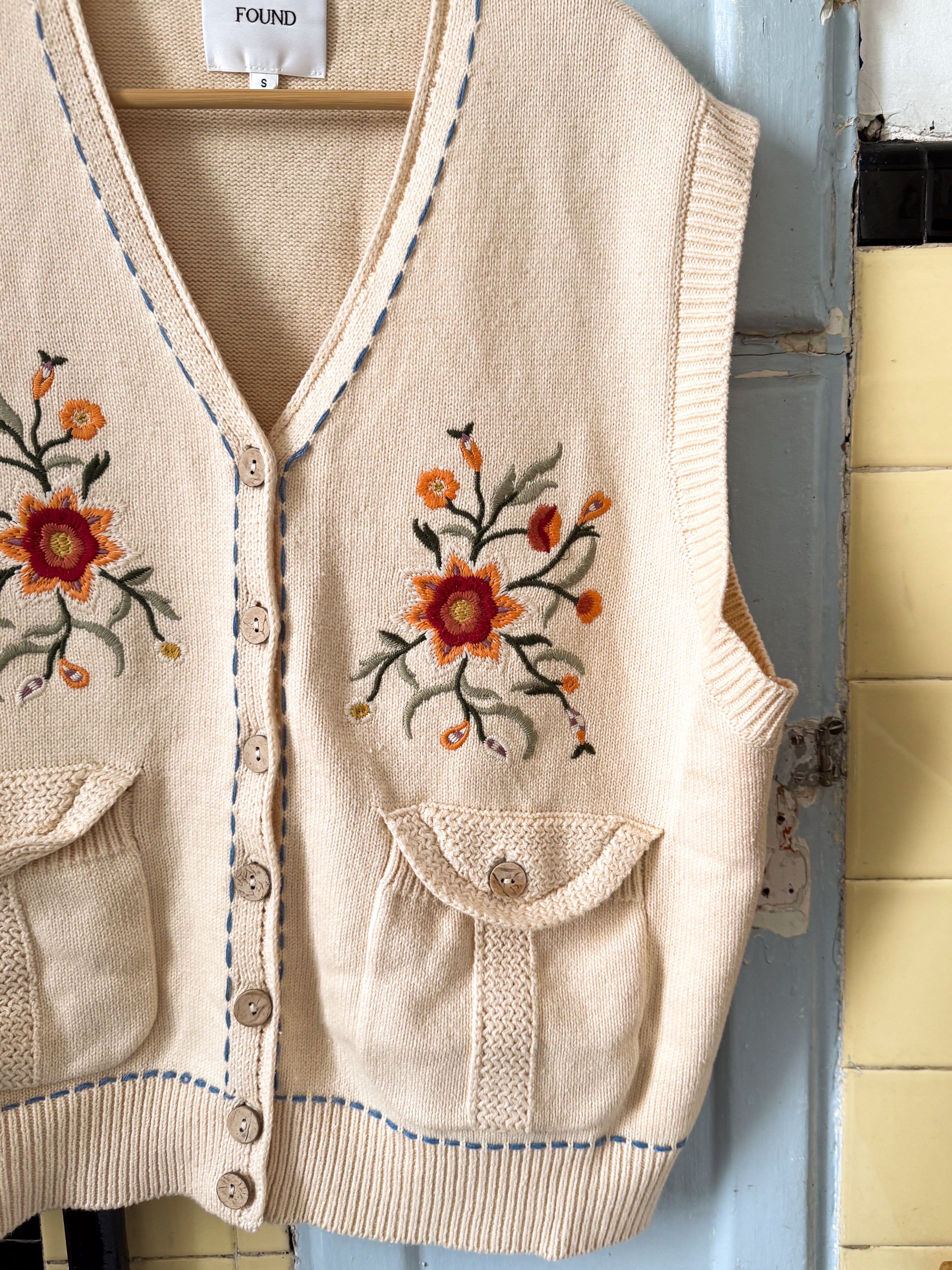 The Vintage-inspired Floral Embroidered Knit Vest by FOUND, cream-colored with red and orange flowers, buttons, and two pockets, hangs against a blue and yellow tiled wall. Ideal for adding unisex charm to any outfit.