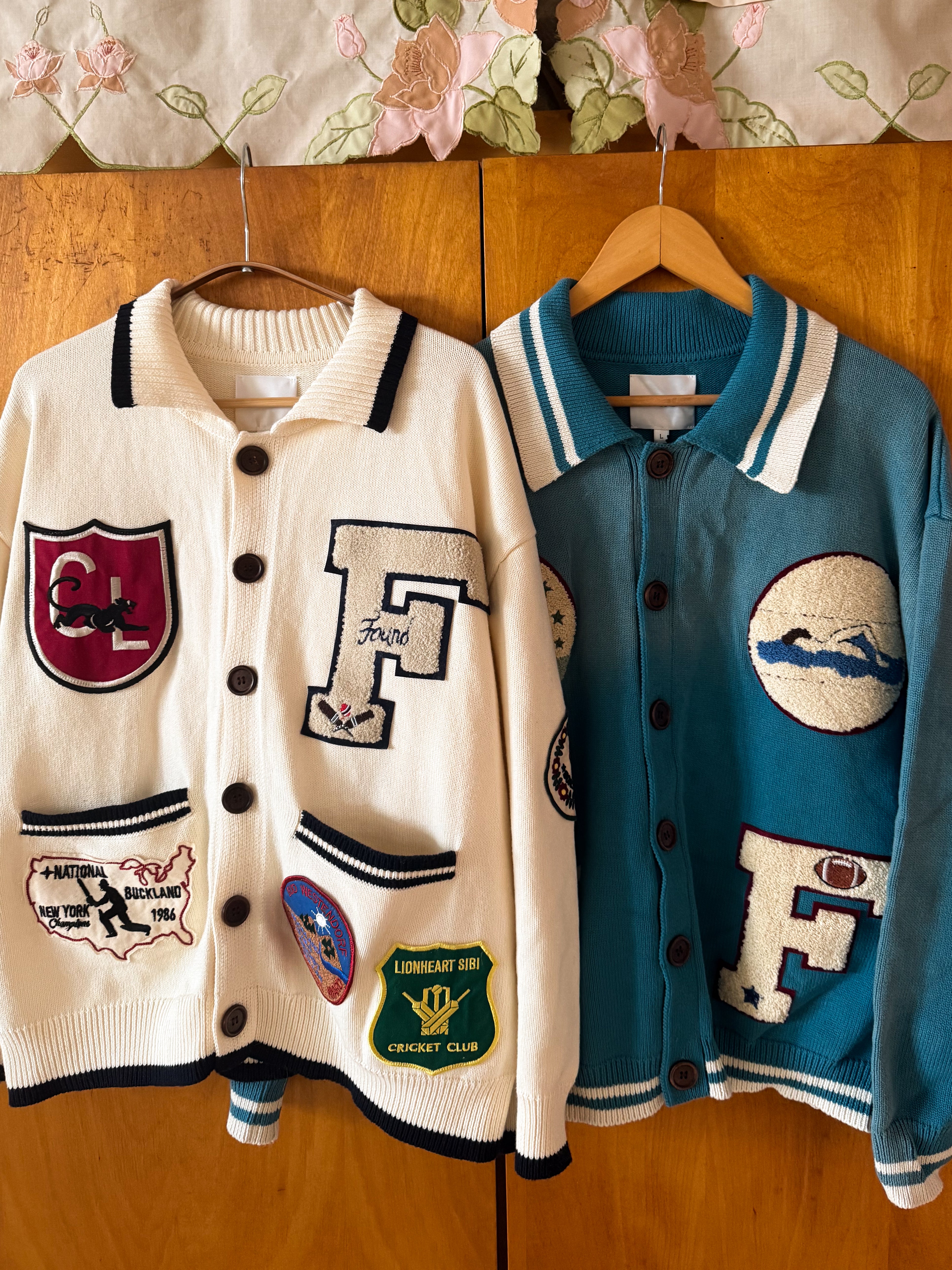 The Fin Varsity Patch Collared Cardigans by FOUND feature a white design with collegiate patches, including a red emblem and green cricket club badge, and a blue version adorned with chenille appliqués like an F and mountain scene for the ultimate varsity vibe.