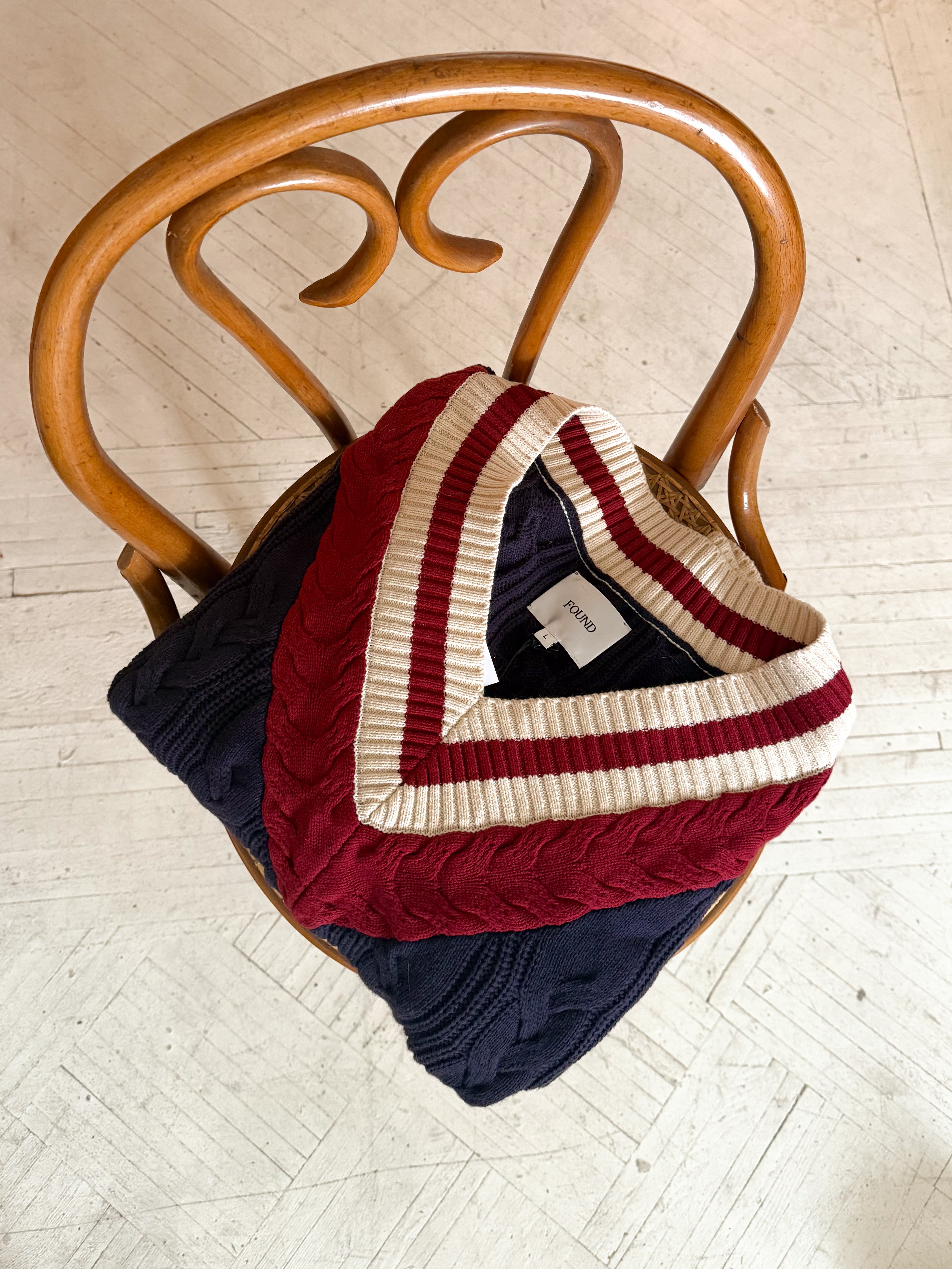 Two folded sweaters, a Found navy cable-knit and a red FOUND V-Neck Cricket Sweater featuring a white-striped collar, elegantly rest on a curved wooden chair.