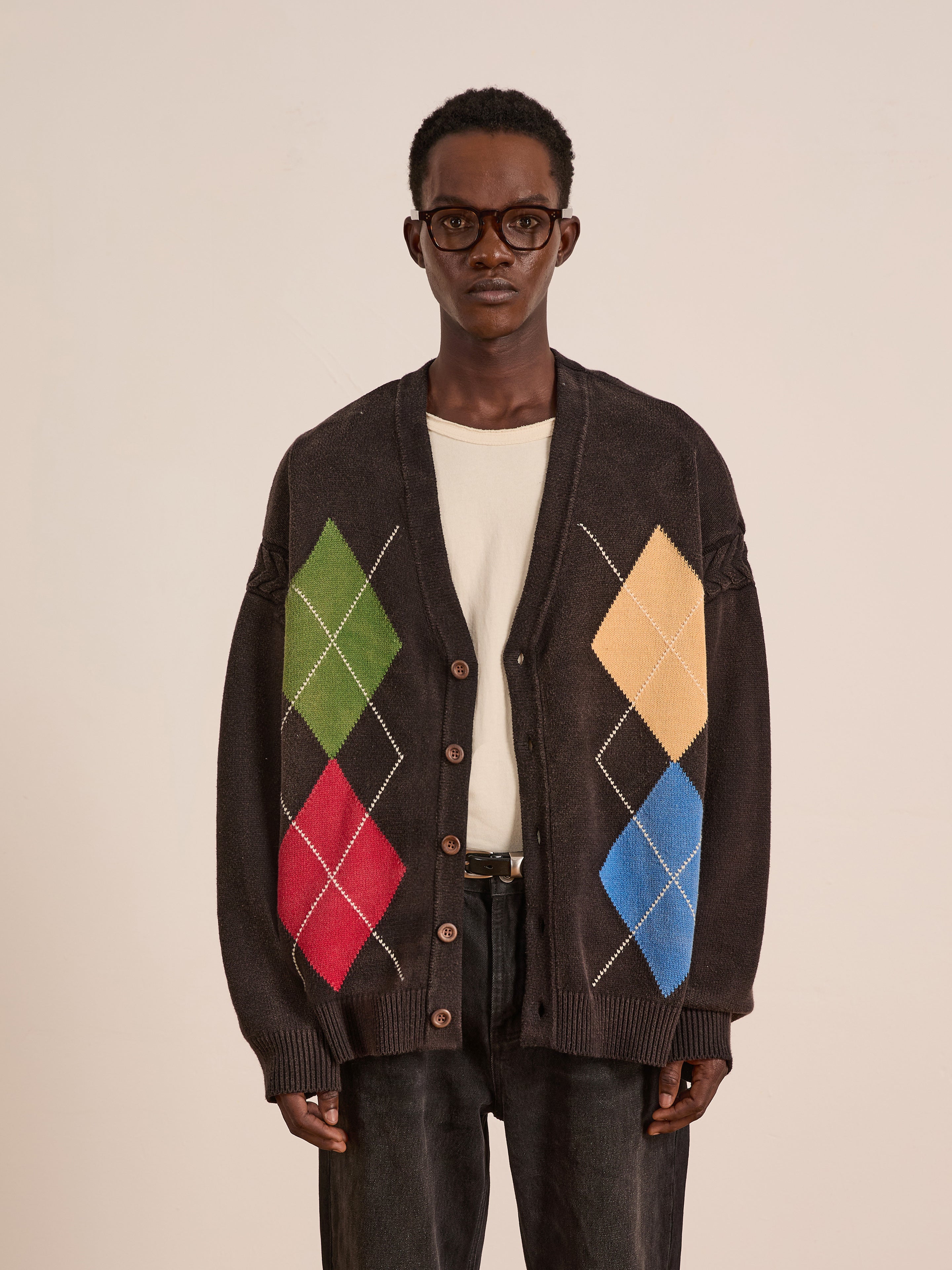 A man in glasses, a cream shirt, and the FOUND Argyle Knit Cardigan stands against a plain background. This unisex piece features bold green, red, yellow, and blue argyle patterns for a standout statement look.